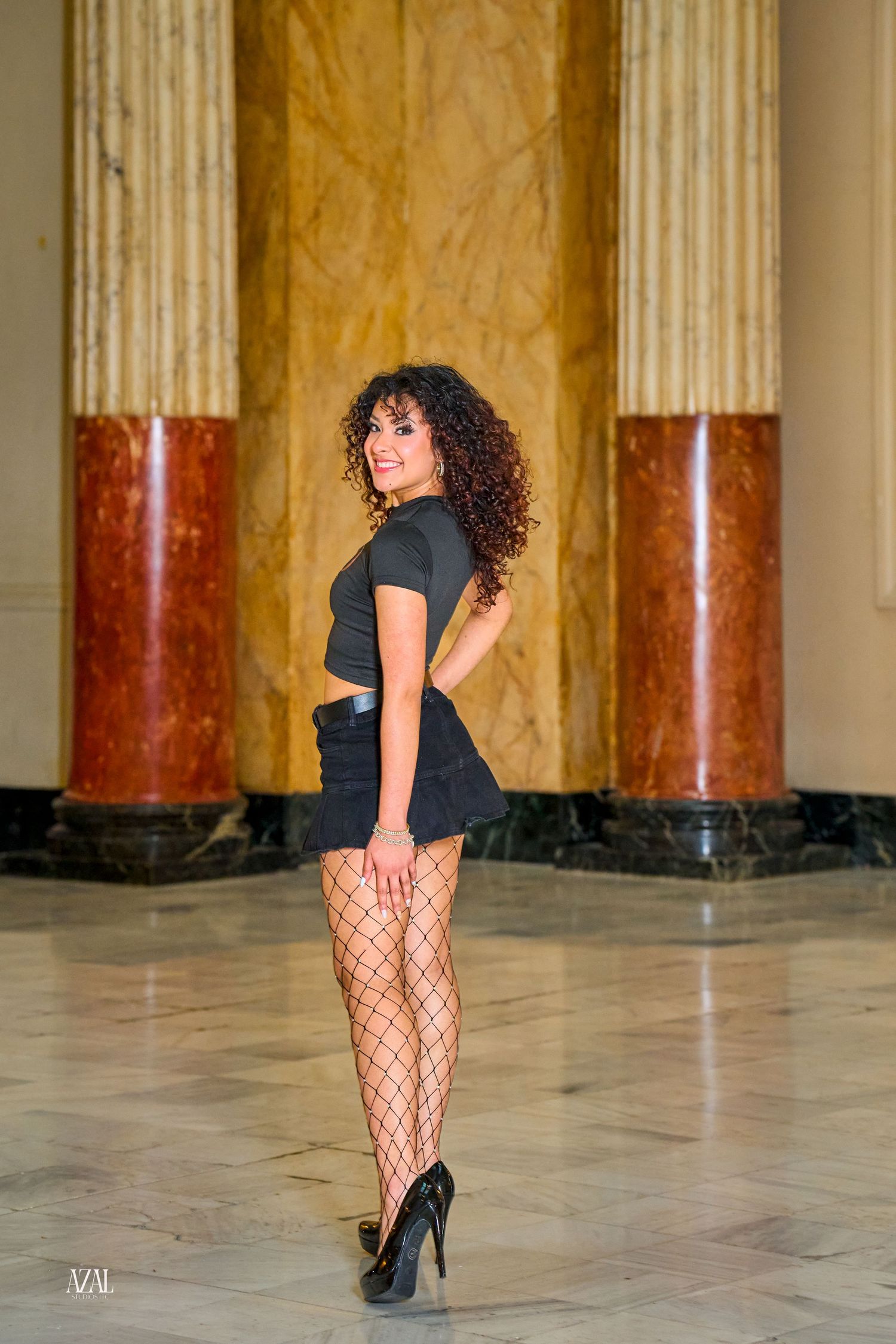 Woman in black mini skirt, fishnet stockings, and heels poses elegantly among marble columns in grand hall.