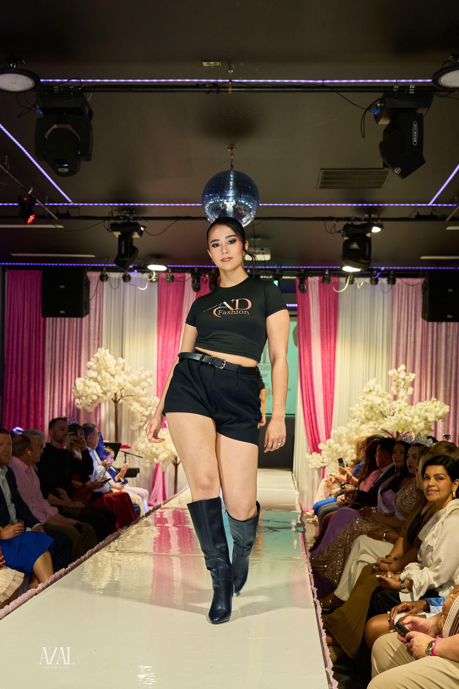 Model in AD Fashion black crop top, shorts, and knee-high boots on lit runway.