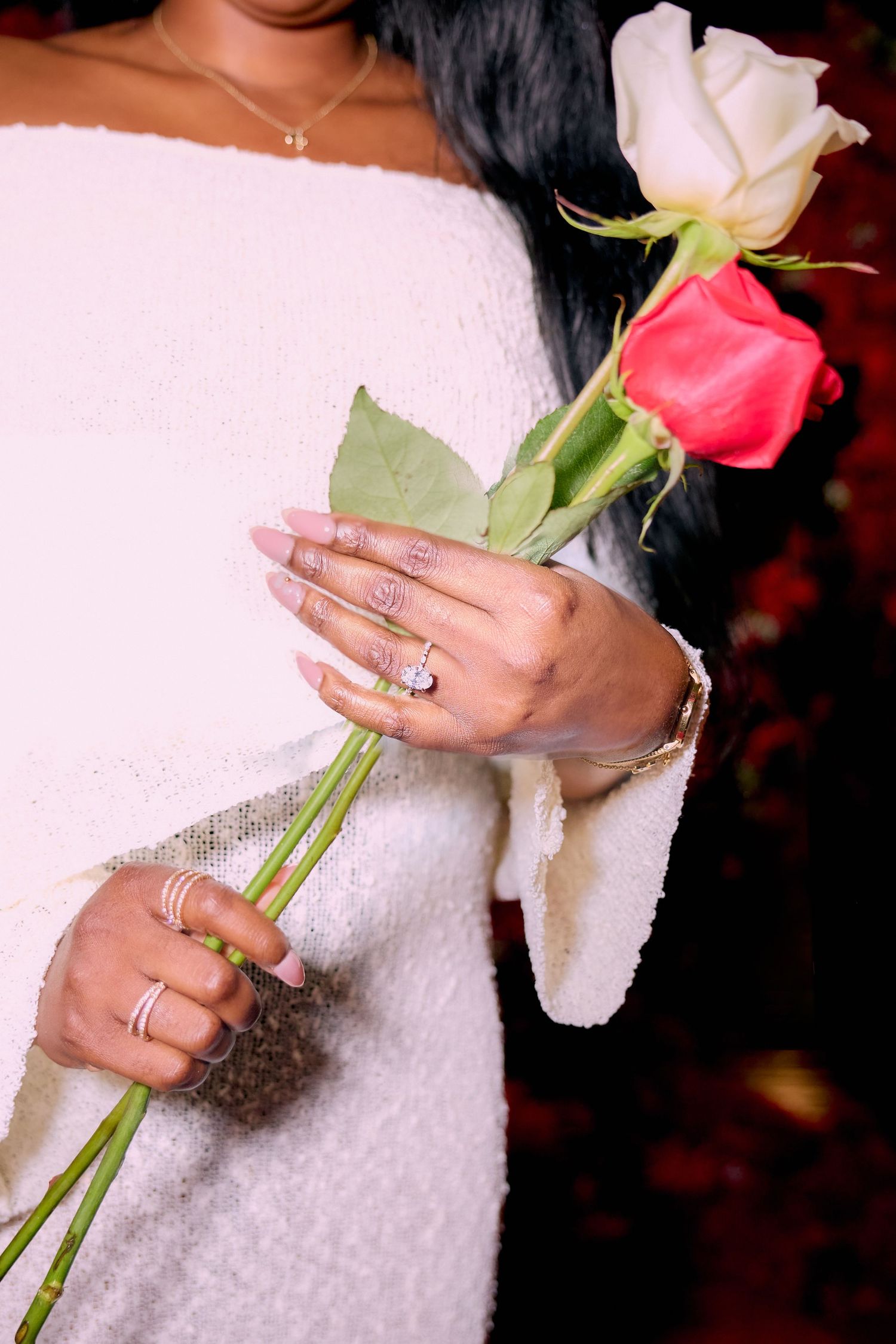 A person in a white dress holds two roses - one pink and one white - while showing off elegant rings and jewelry.