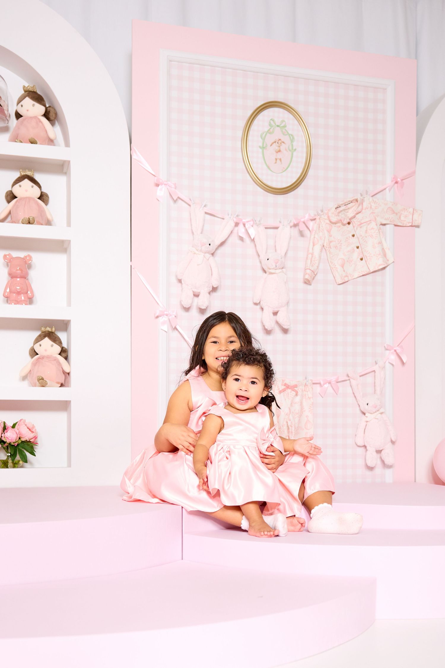 Two young girls in pink dresses pose together in a charming pink nursery-themed setting with bunny decorations.