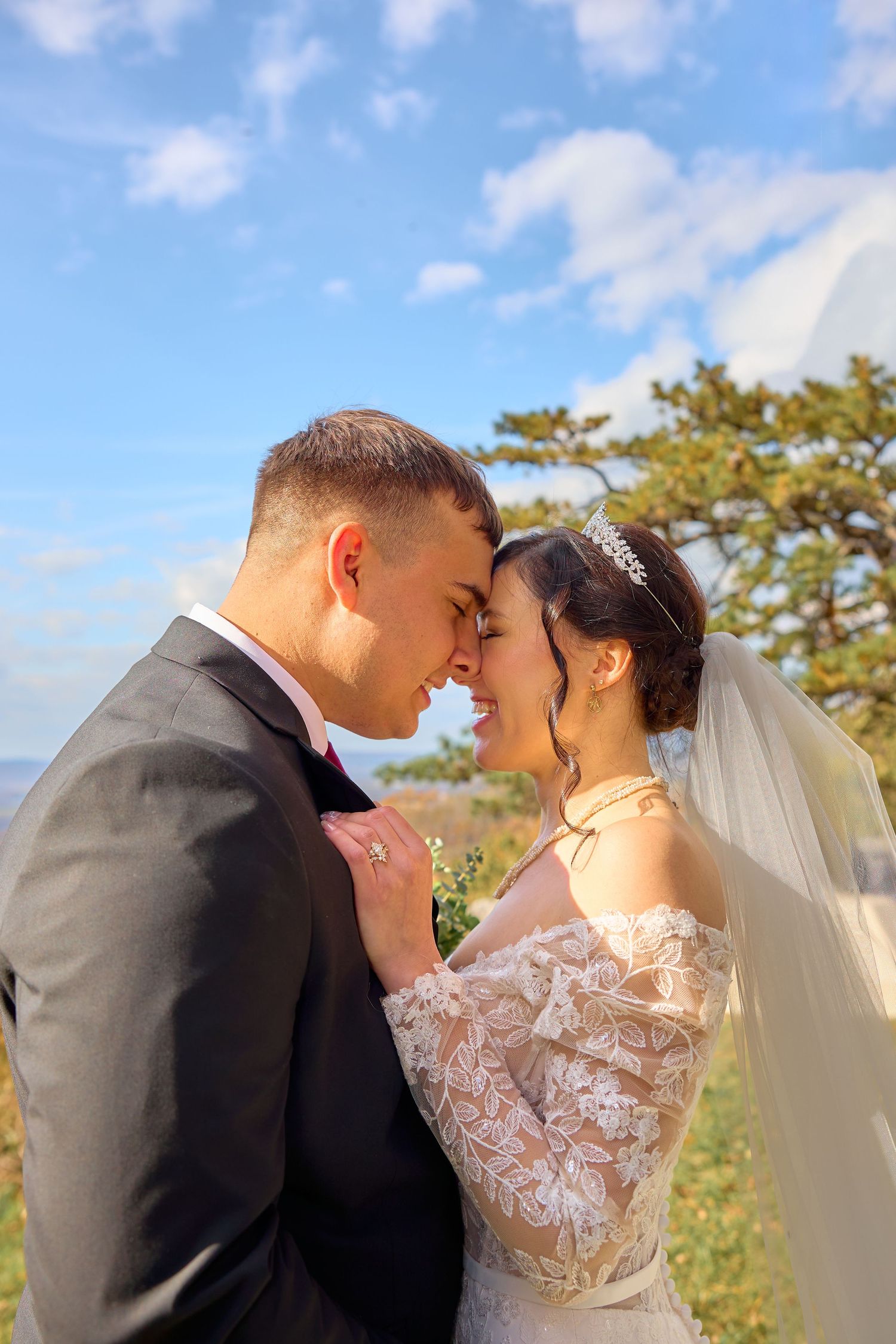 A romantic wedding couple shares an intimate moment outdoors with blue skies and trees in the background.