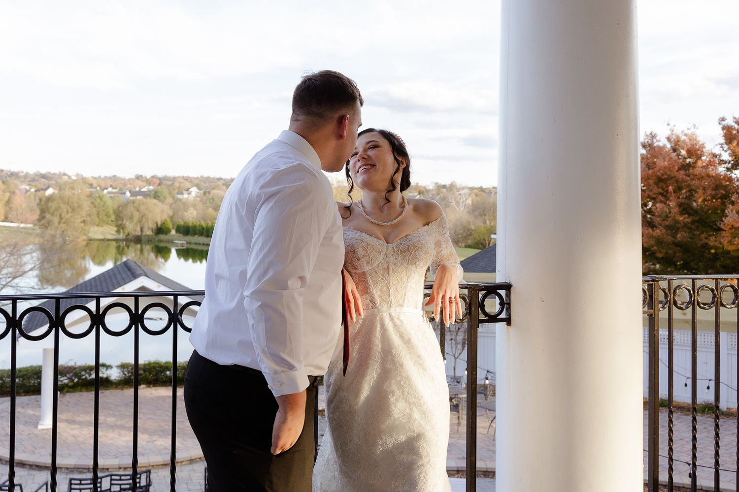 A couple poses romantically on a waterfront terrace with white columns and an ornate railing in the background.