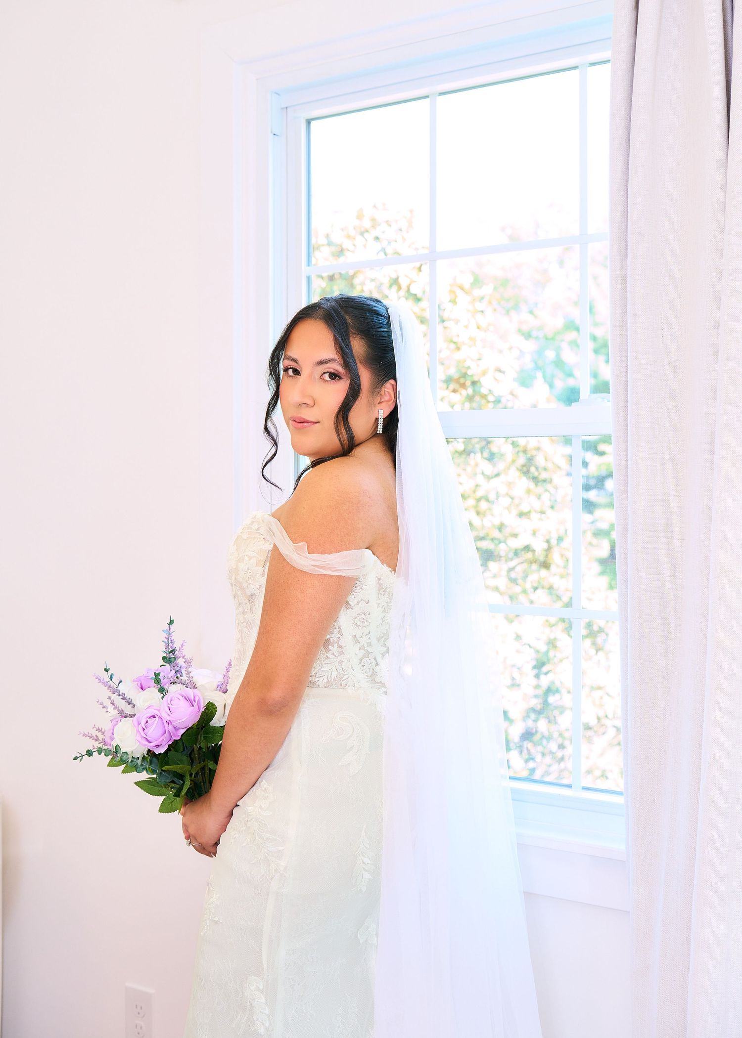 Bride in off-shoulder lace wedding gown and veil holds purple bouquet, posing elegantly by a sunlit window.