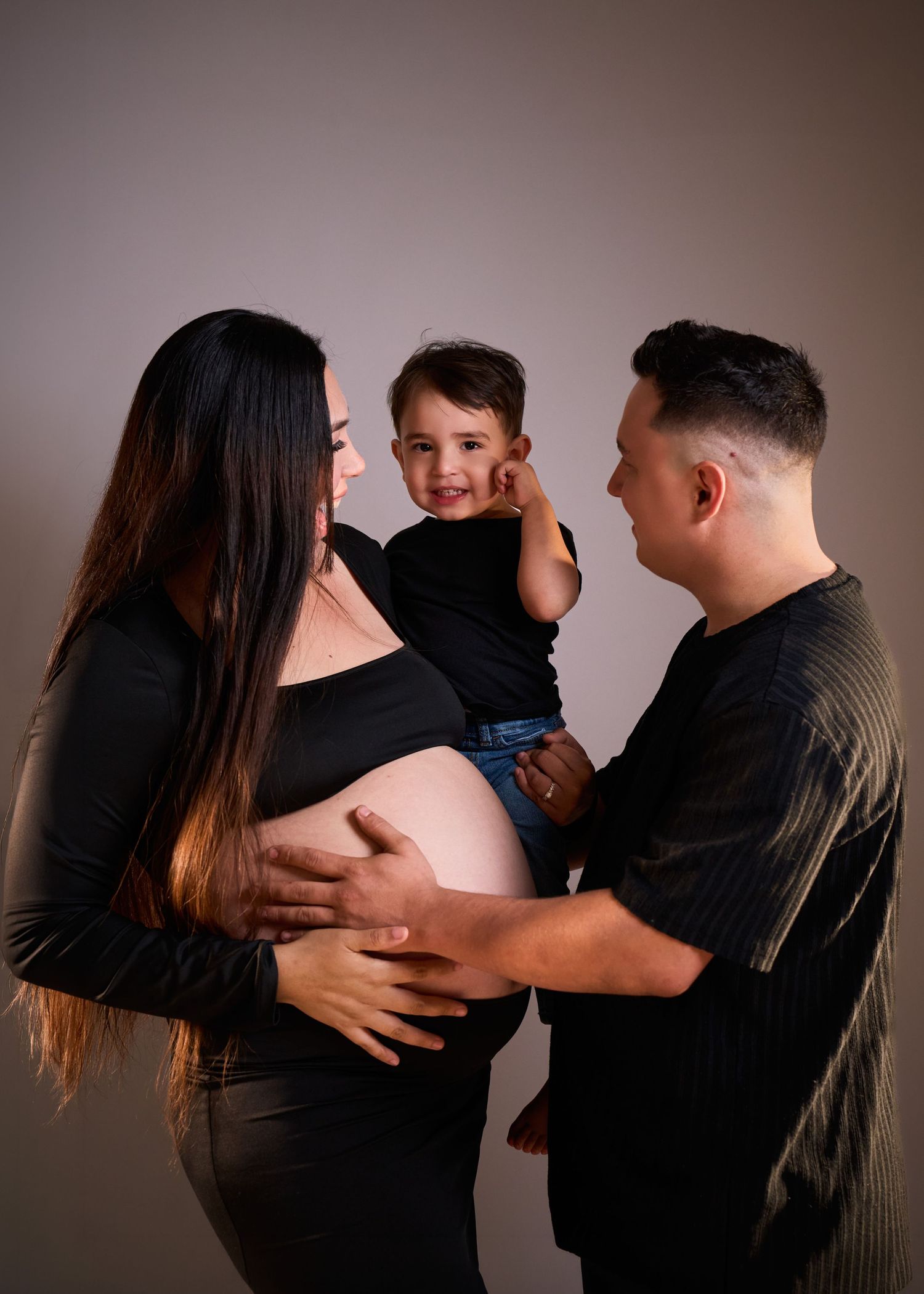 Pregnant woman in black top cradles baby bump while partner holds toddler son in a studio maternity family portrait.