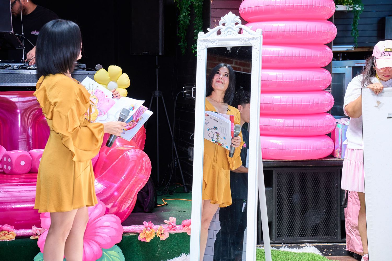 Woman in yellow dress reads magazine reflected in ornate white mirror at pink balloon-decorated outdoor event.