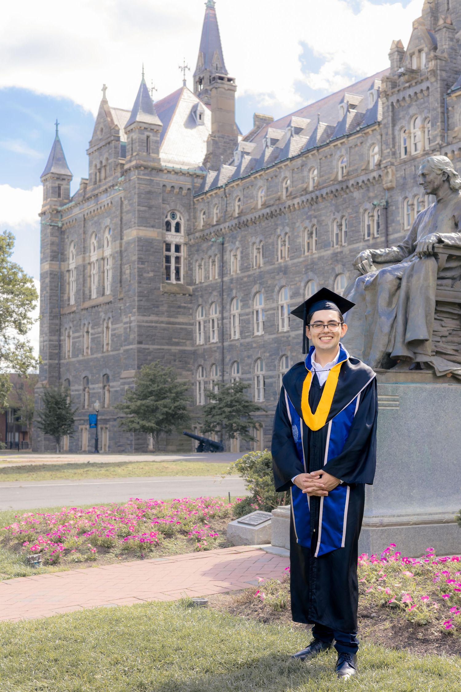 Graduate in blue and gold regalia poses in front of a Gothic university building on graduation day.