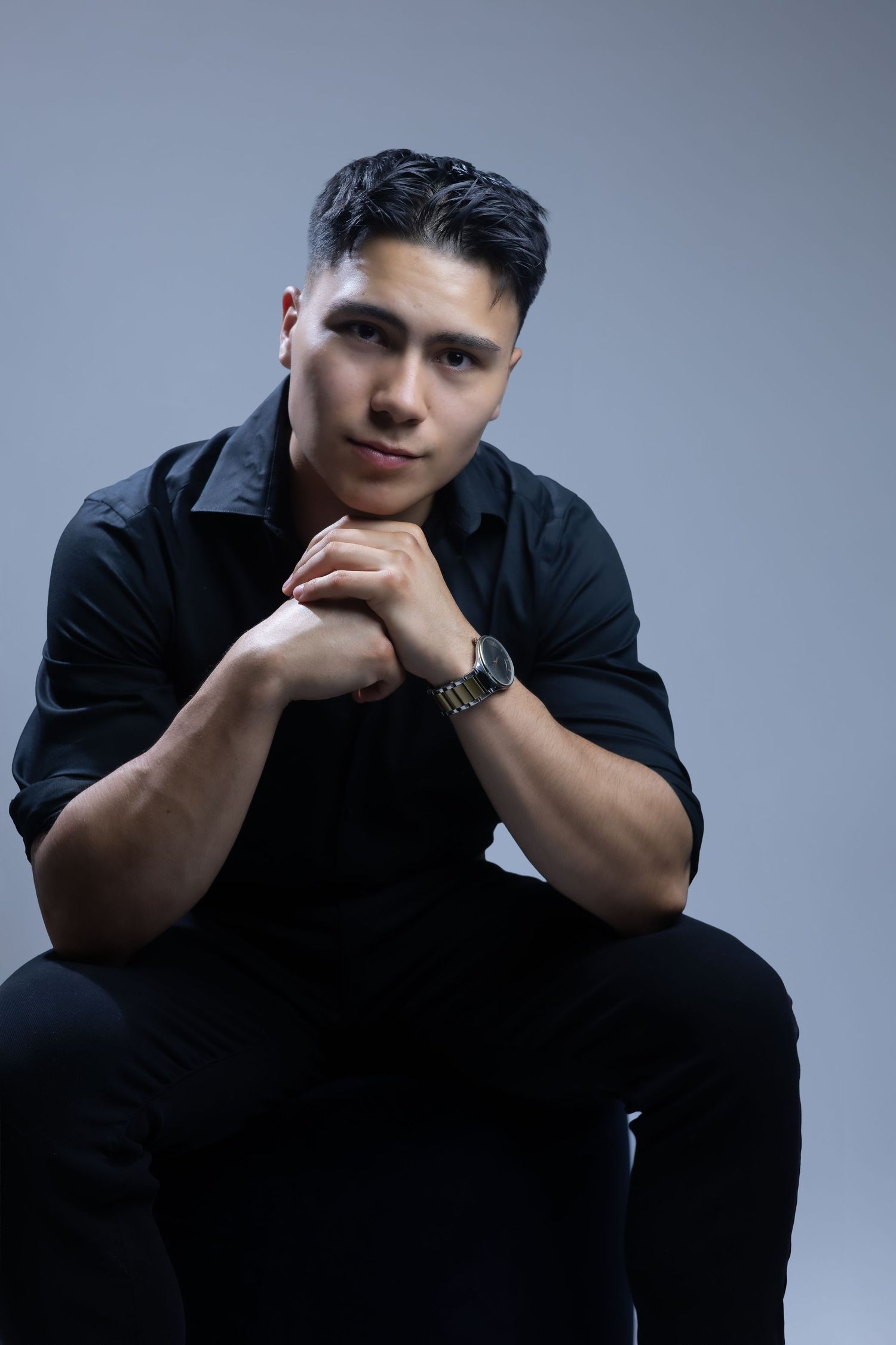 Young model in black attire poses thoughtfully with clasped hands while sitting against gray studio backdrop.