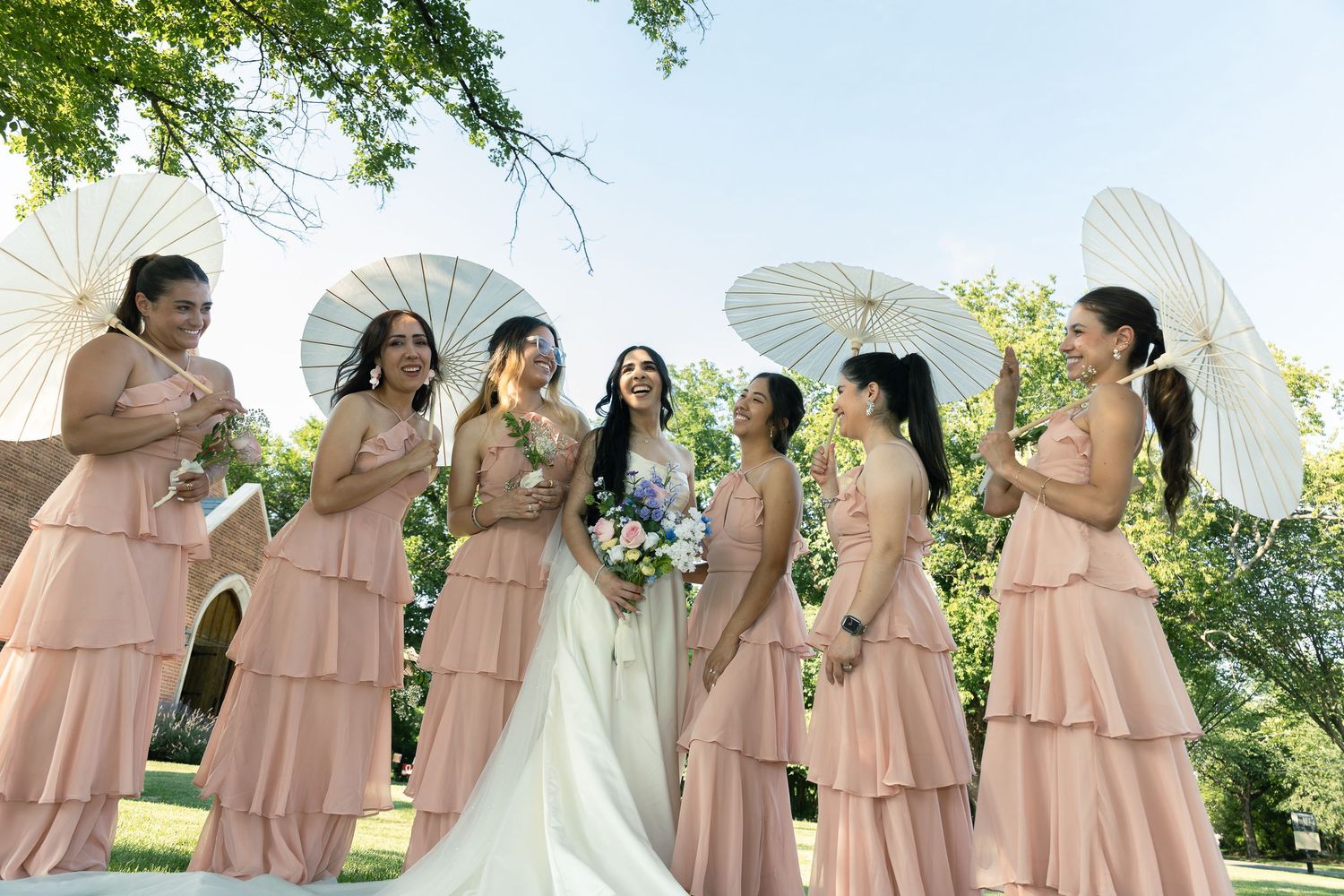 Bridesmaids in blush pink dresses stand with parasols next to bride in white gown during outdoor summer wedding ceremony.