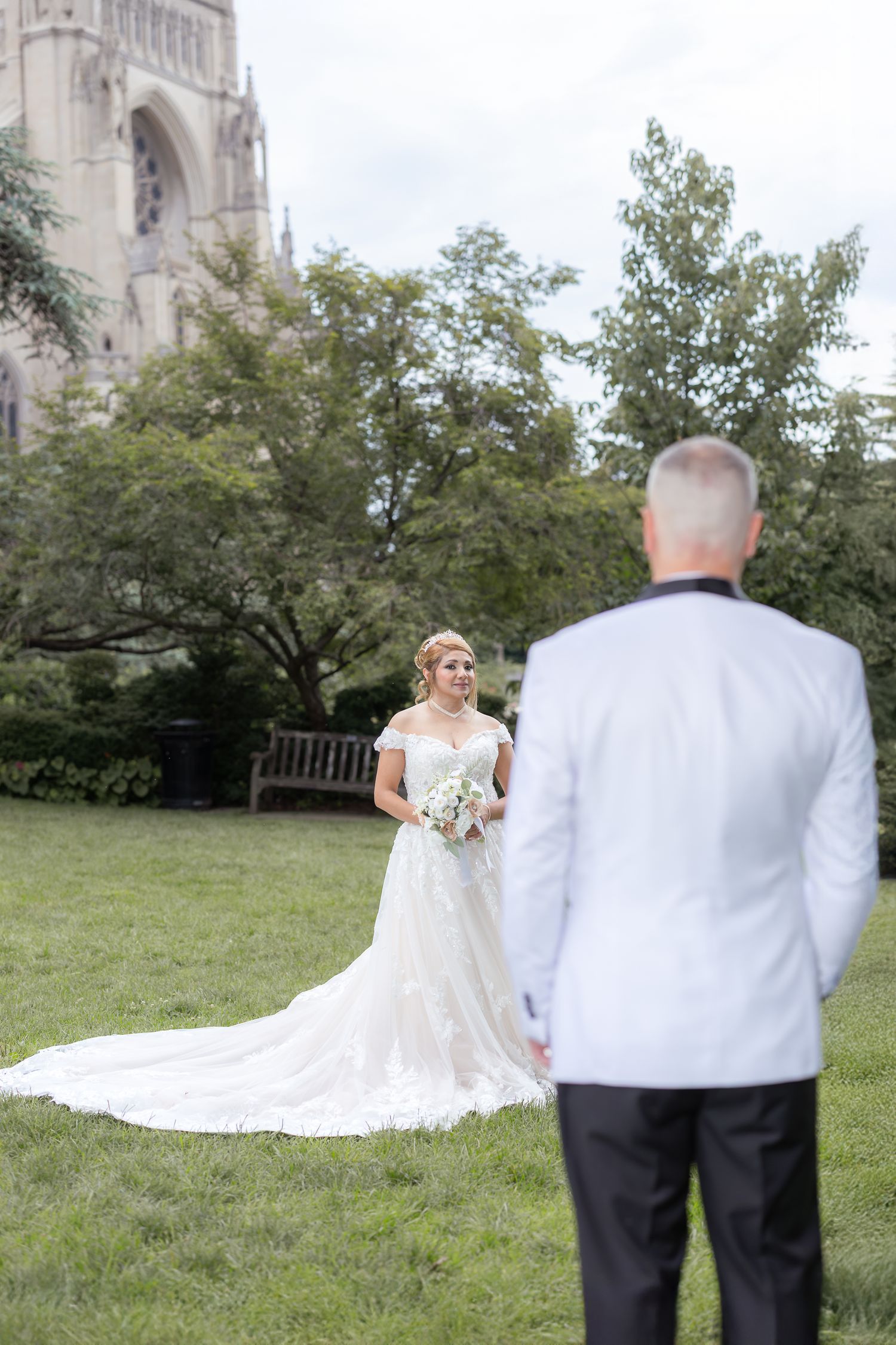 A bride in a flowing white wedding dress stands on grass outside a church while looking at a person wearing a white suit.