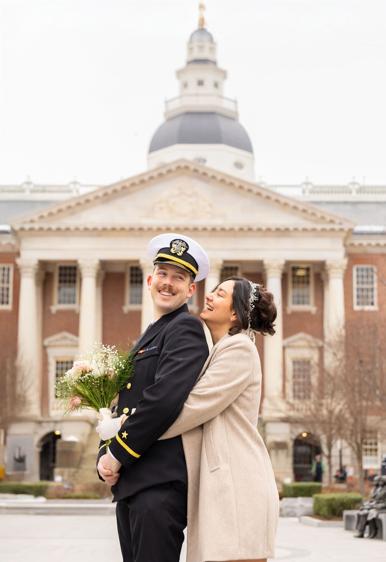 Couple embraces in front of historic government building with white columns and dome.
