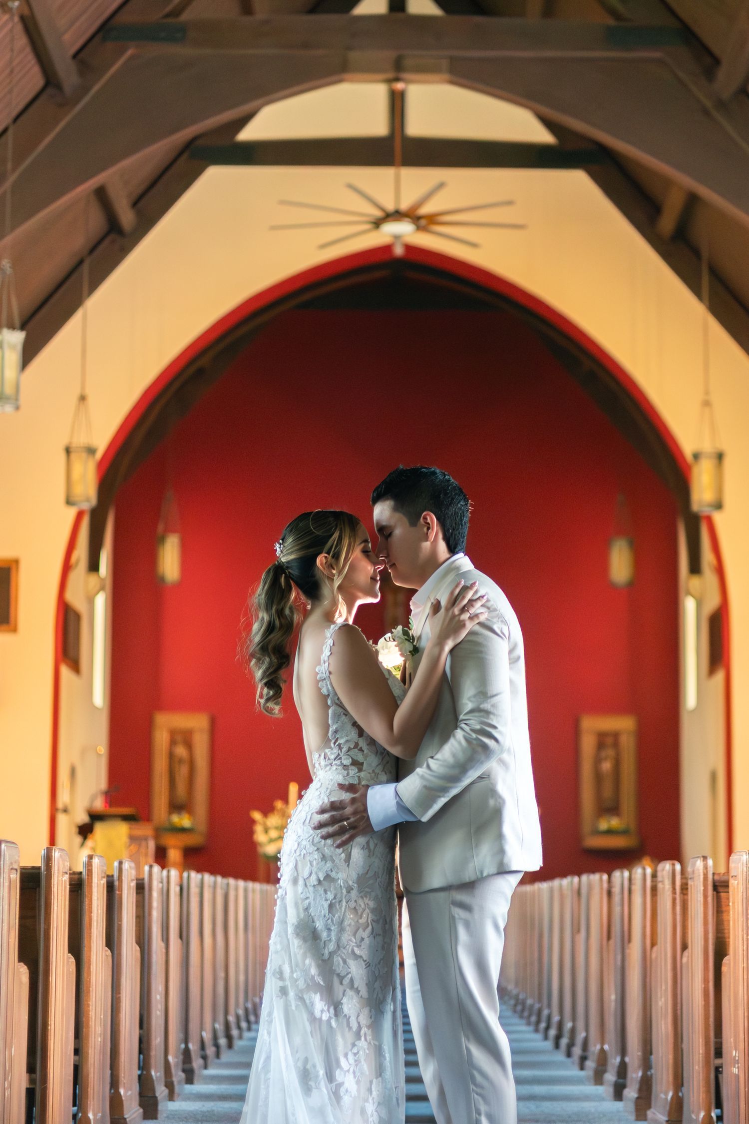 A romantic wedding couple stands in an elegant church hallway with red walls and arched wooden beams.