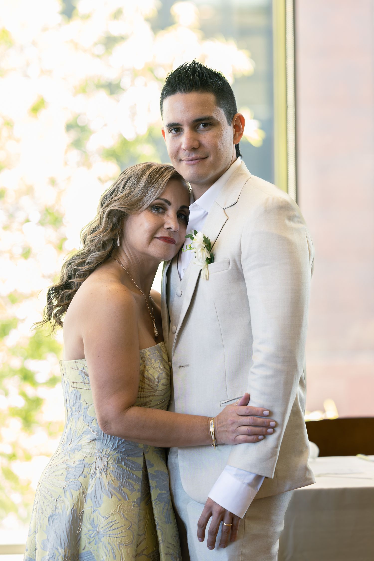 Couple in white formal attire pose together near bright windows.