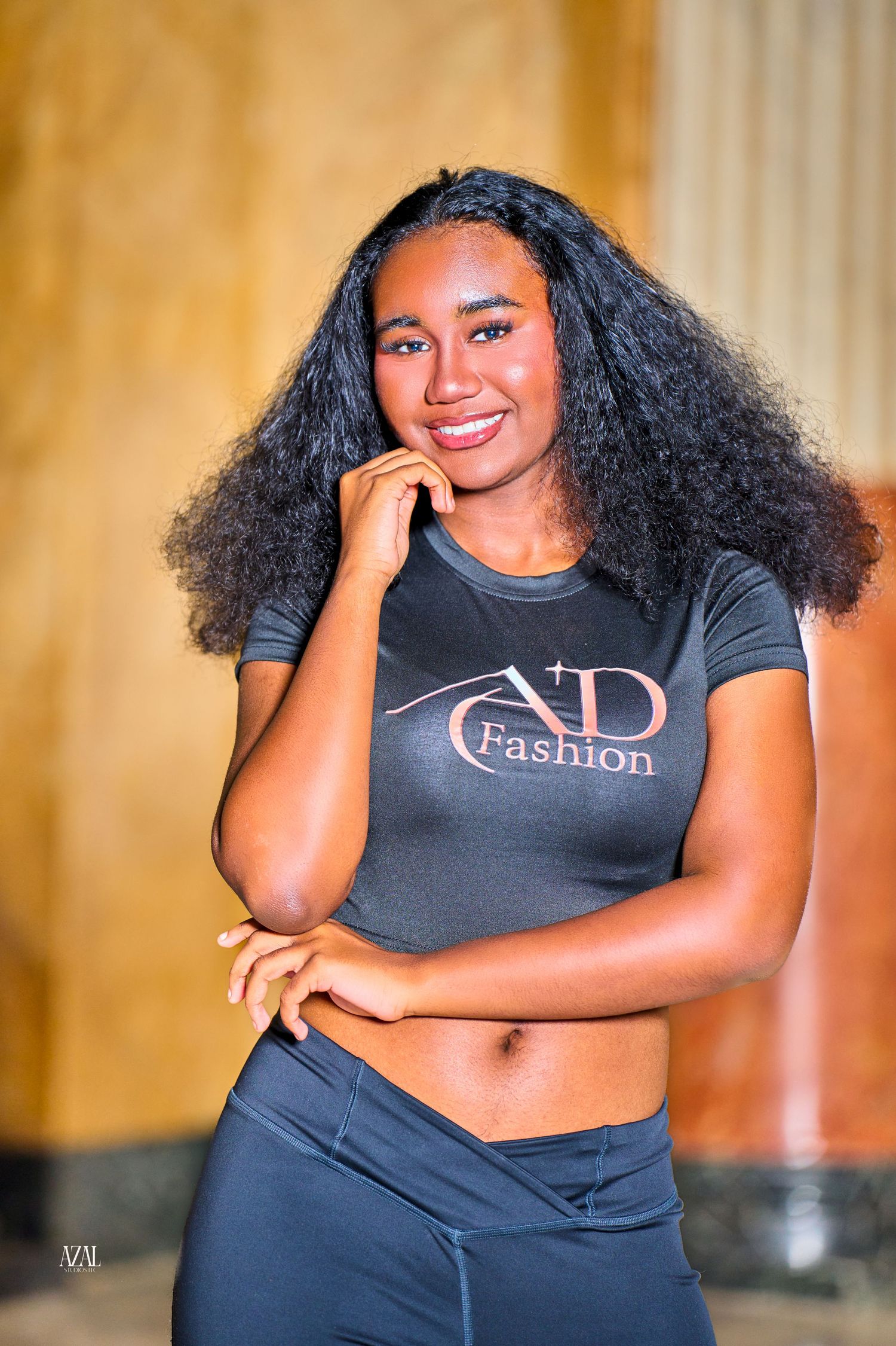 Smiling young woman with curly hair wearing an AD Fashion black crop top and leggings poses elegantly.