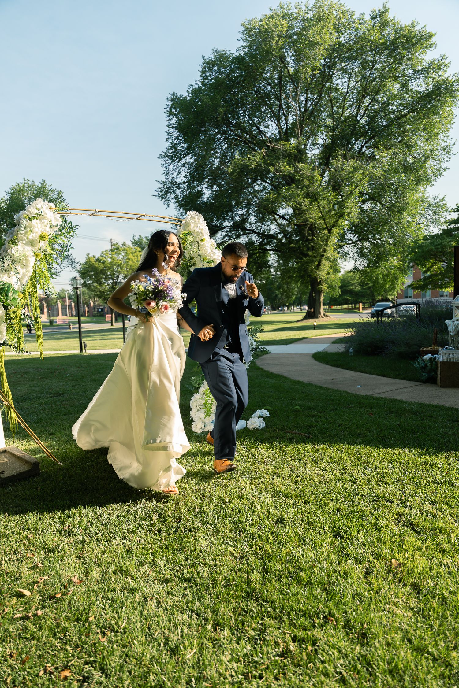 A couple walks together on a grass lawn under a decorated arch during an outdoor wedding ceremony.