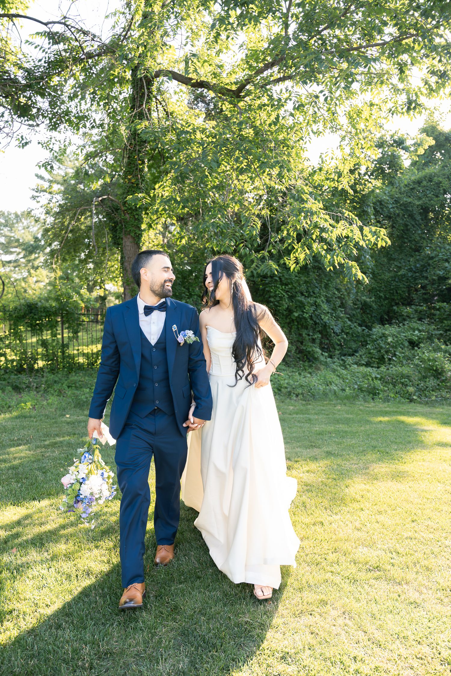 A newlywed couple walks on a sunlit lawn with green trees in the background during their summer outdoor wedding.
