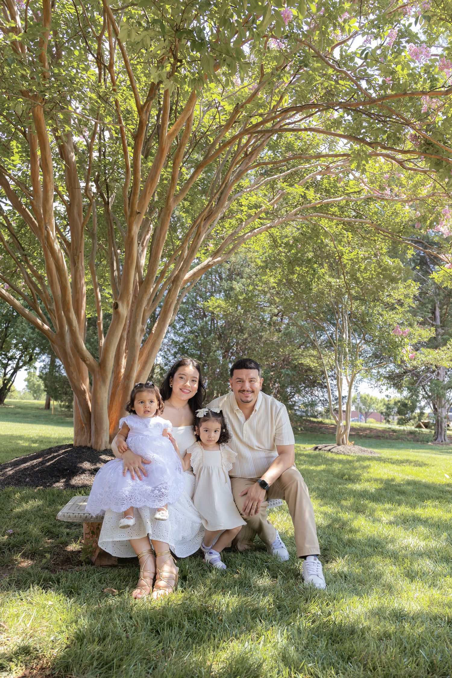 Family group seated under large tree wearing coordinated white outfits for outdoor photo session.