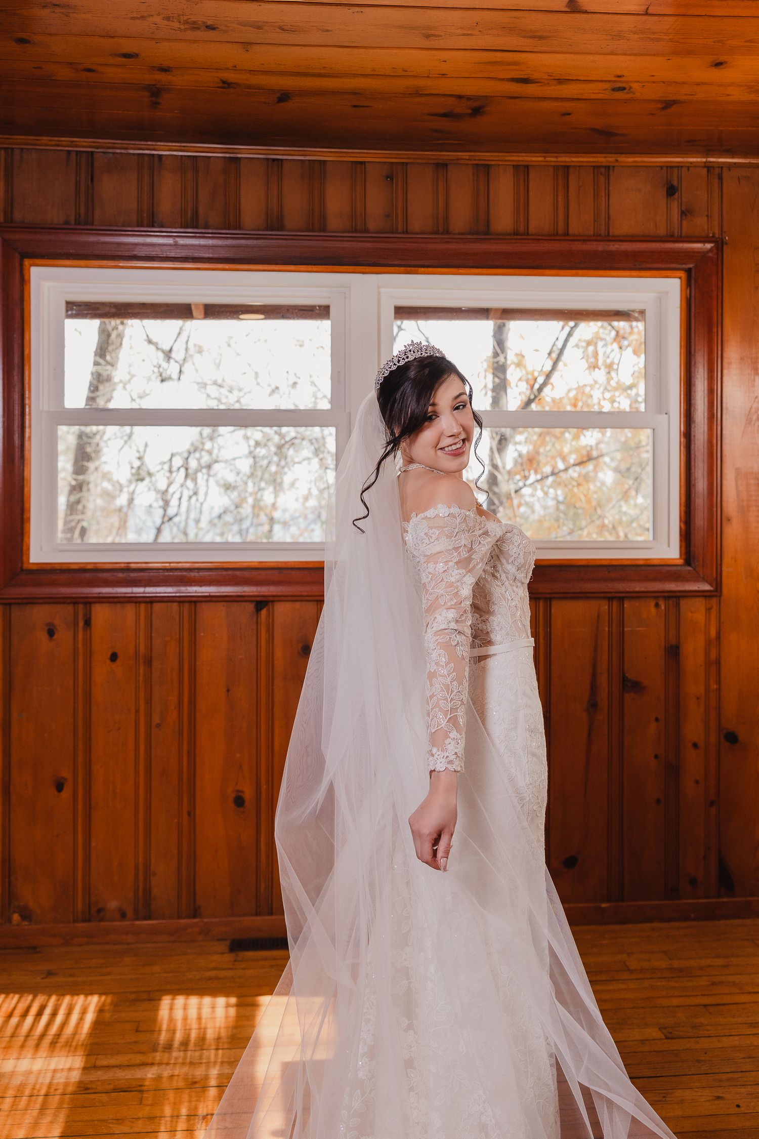 A flowing white wedding veil trails behind in a warm wood-paneled room with natural lighting.