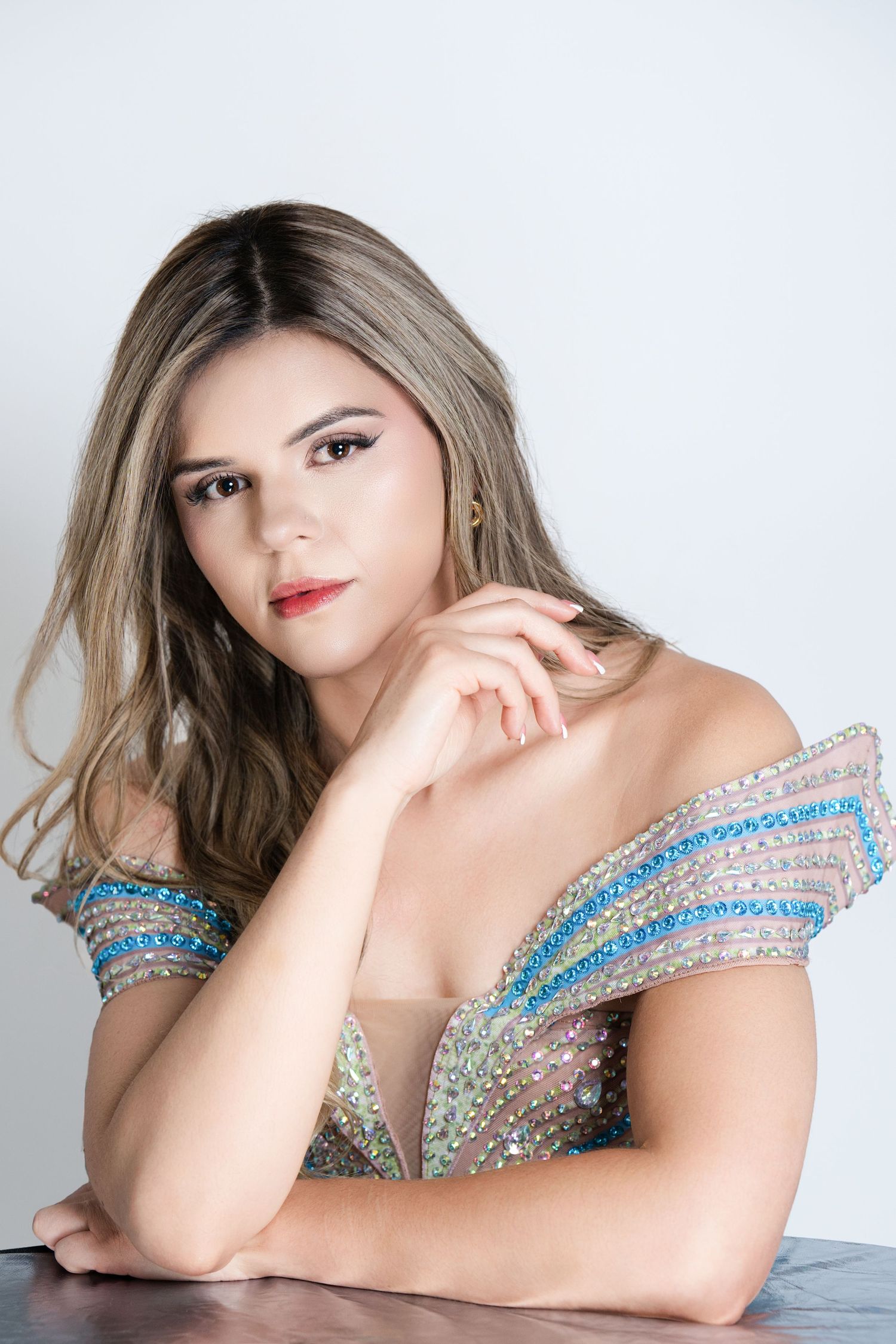 Young woman in sequined off-shoulder dress poses elegantly at table against white background.