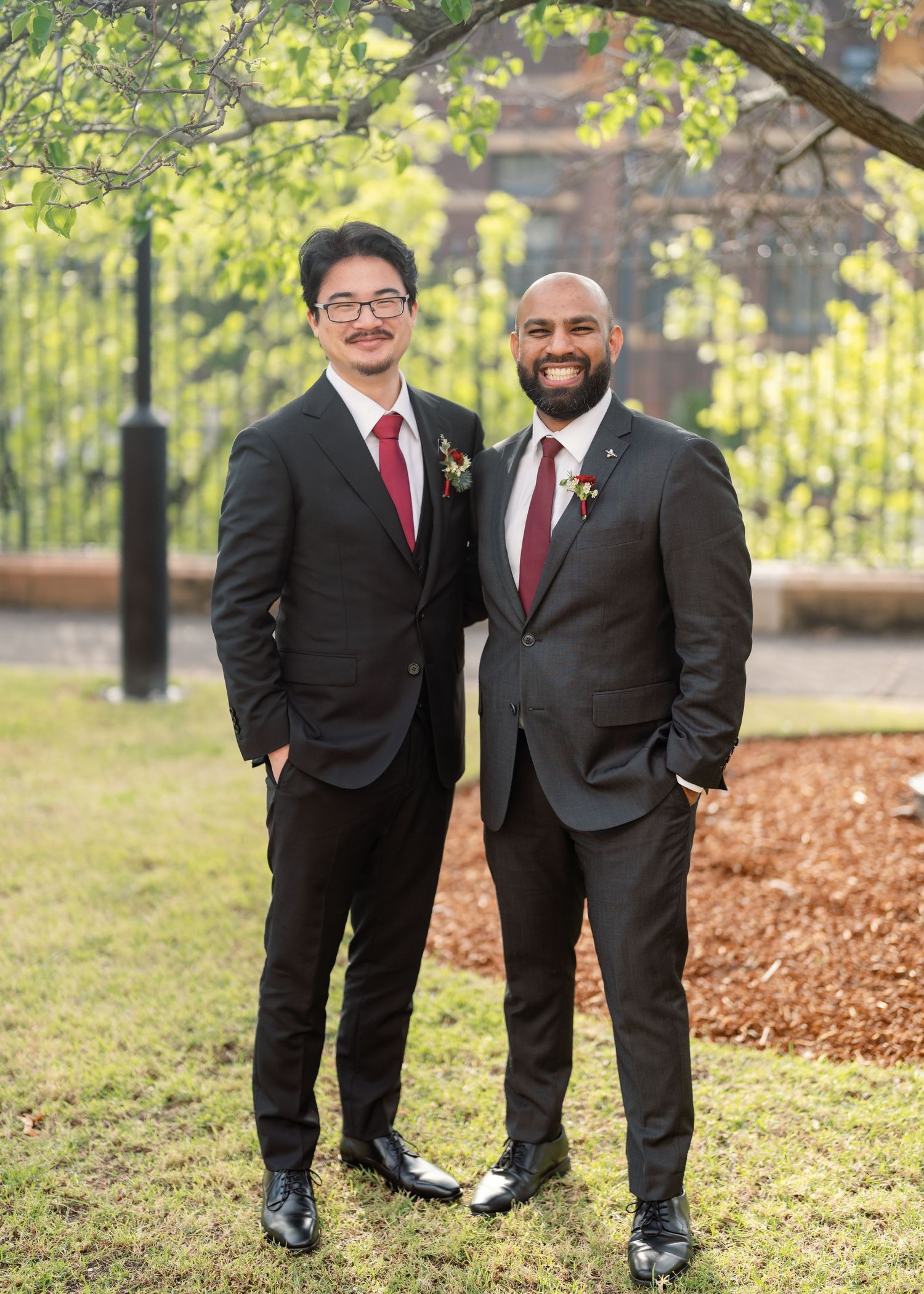 Wedding party members in black suits pose playfully on grass near trees.