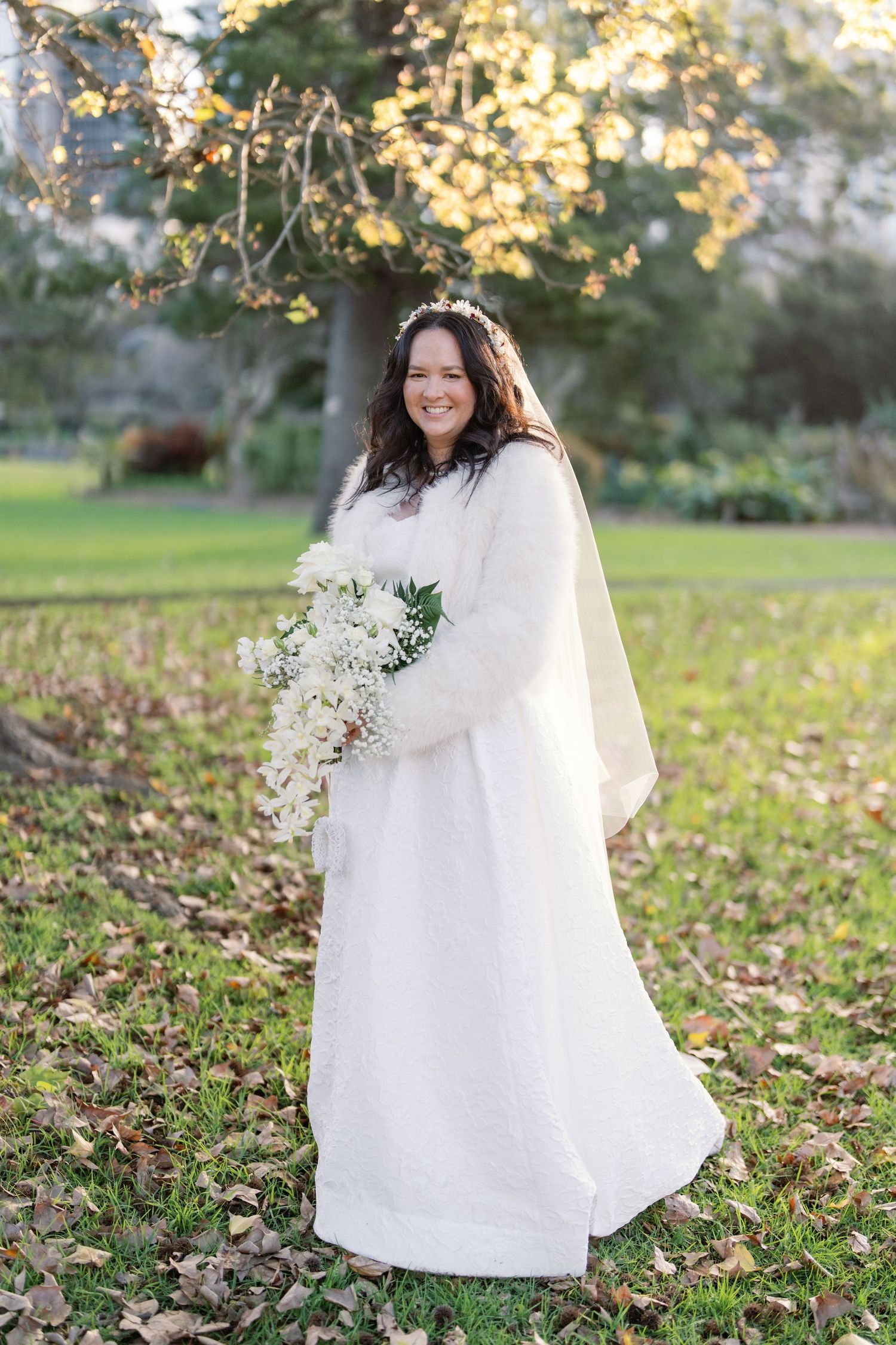 Smiling bride in white lace gown holding cascading white bouquet, wearing floral crown and veil outdoors.