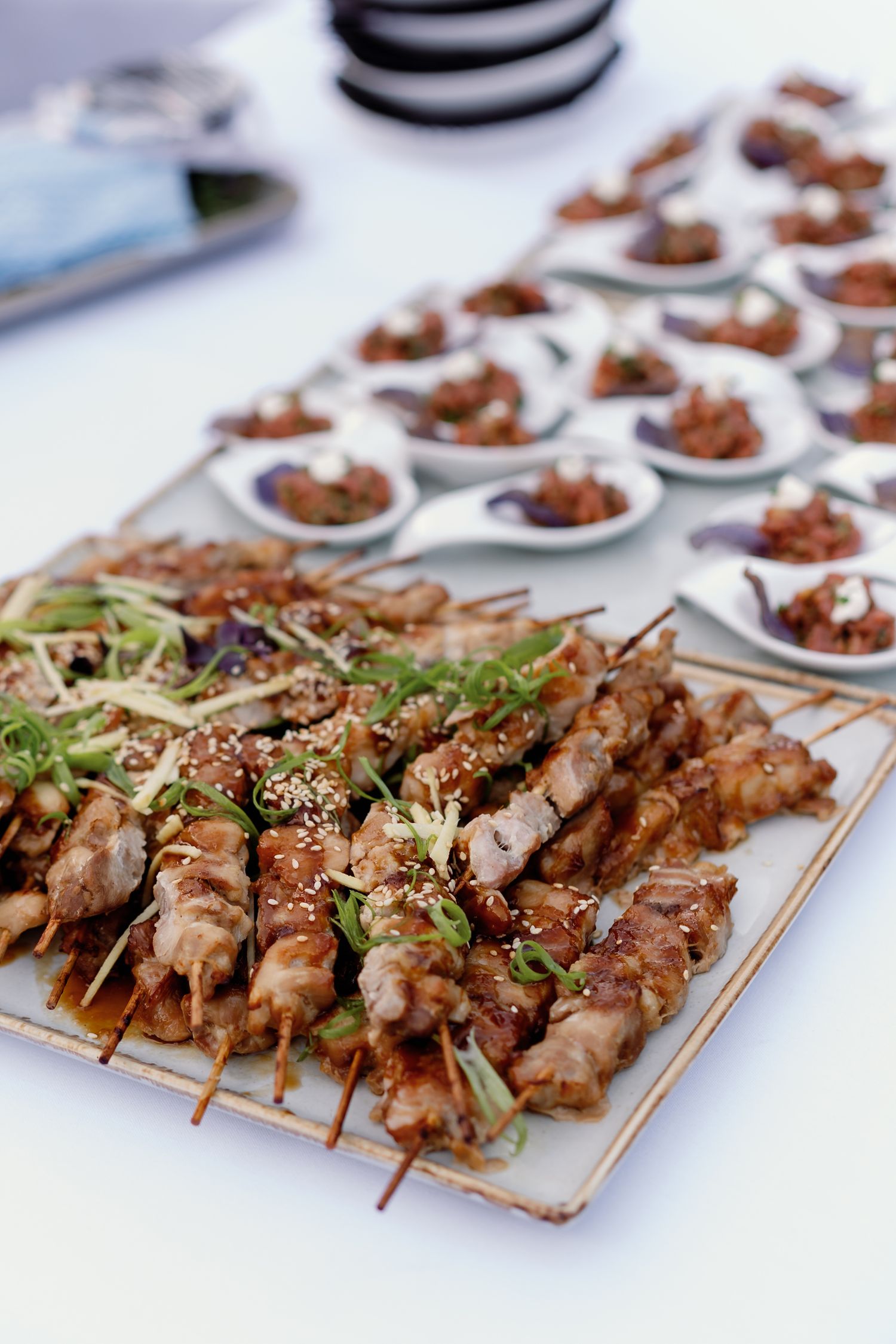 Grilled meat skewers topped with sesame seeds and herbs on a tray, with small appetizer spoons in the background.