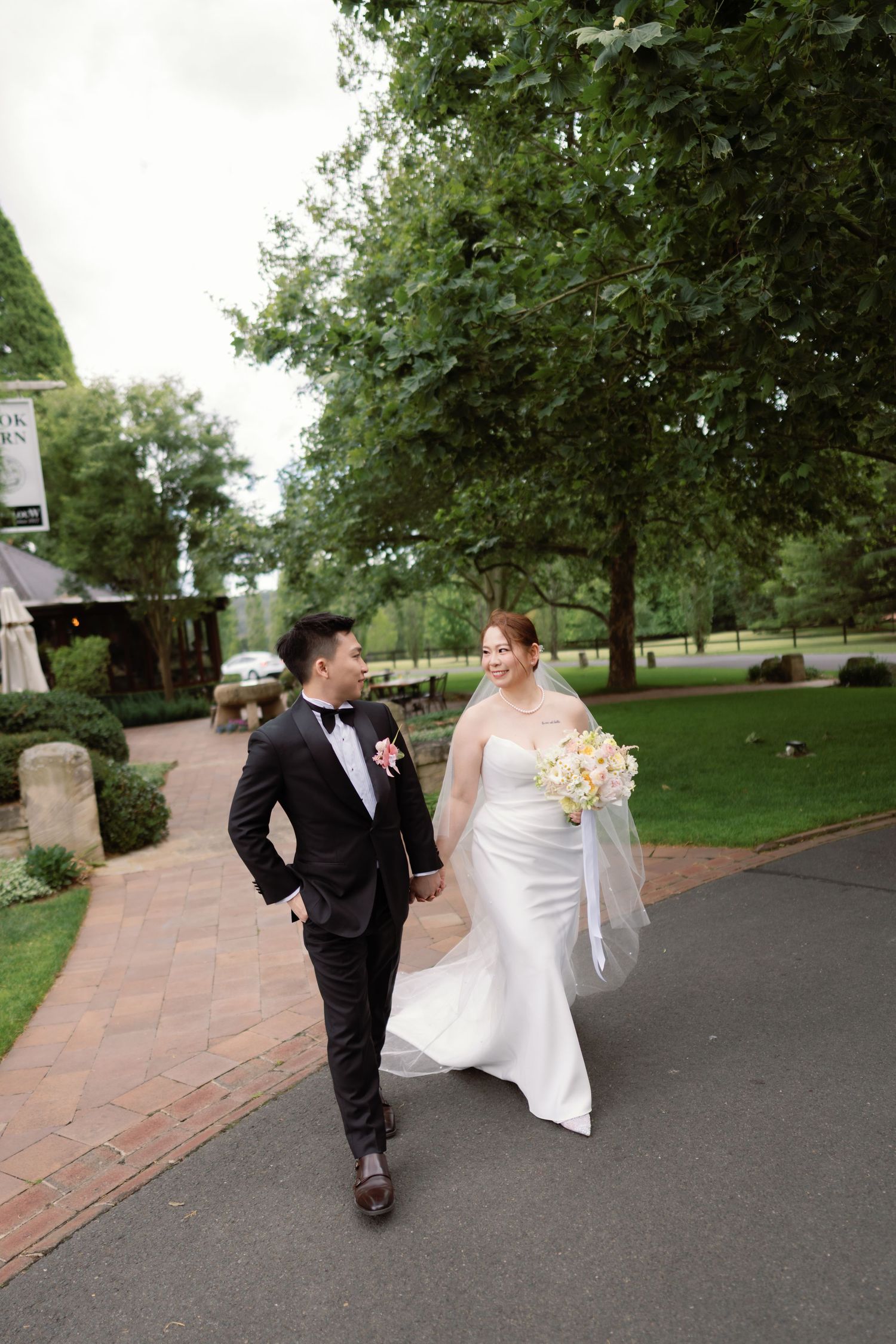 Bride and groom walk hand-in-hand along a brick path surrounded by lush green trees at an elegant outdoor wedding venue.