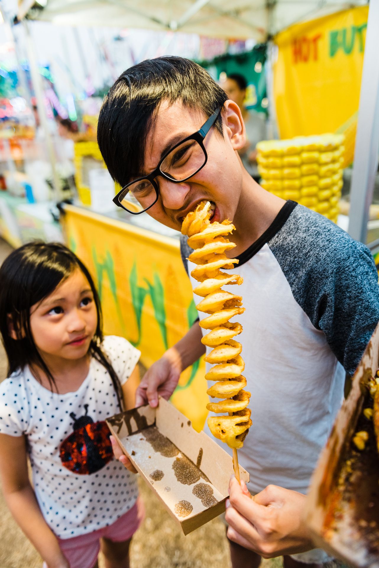 Someone enjoying a unique spiral potato snack on a stick at an Asian street food market while another person watches.