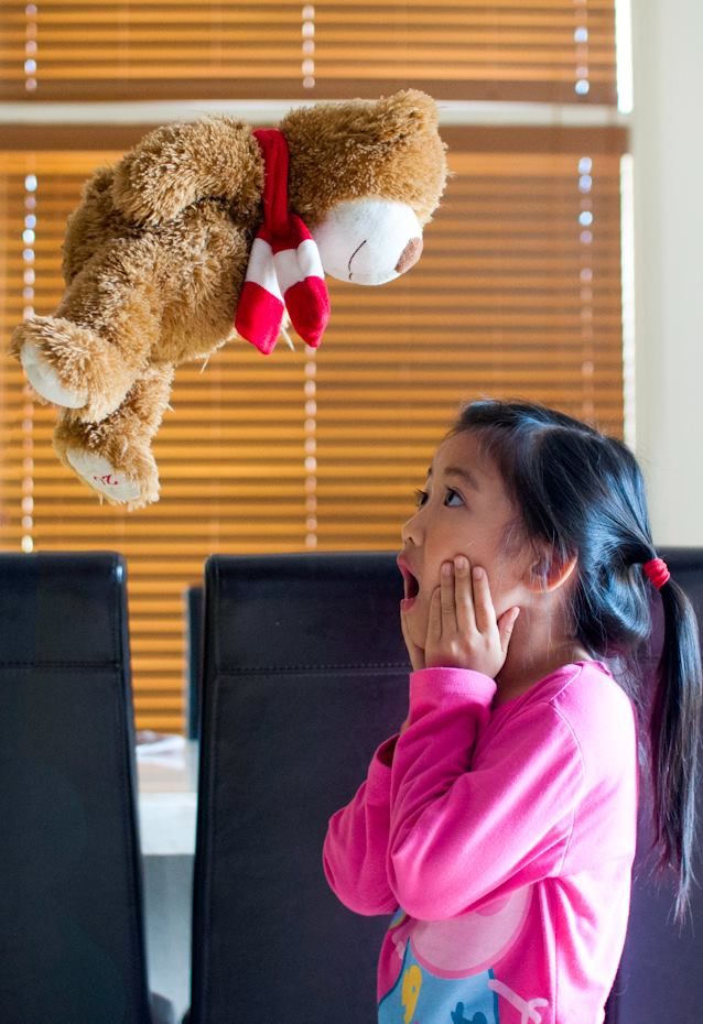 A child looking surprised at a floating teddy bear in front of window blinds.
