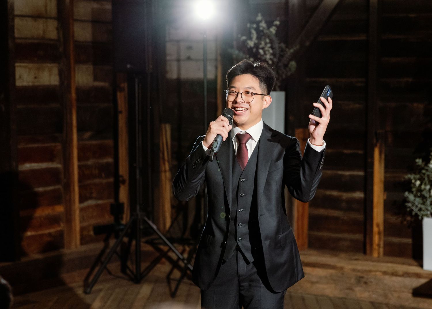 A groom poses in a black suit against a rustic wooden backdrop with dramatic lighting.