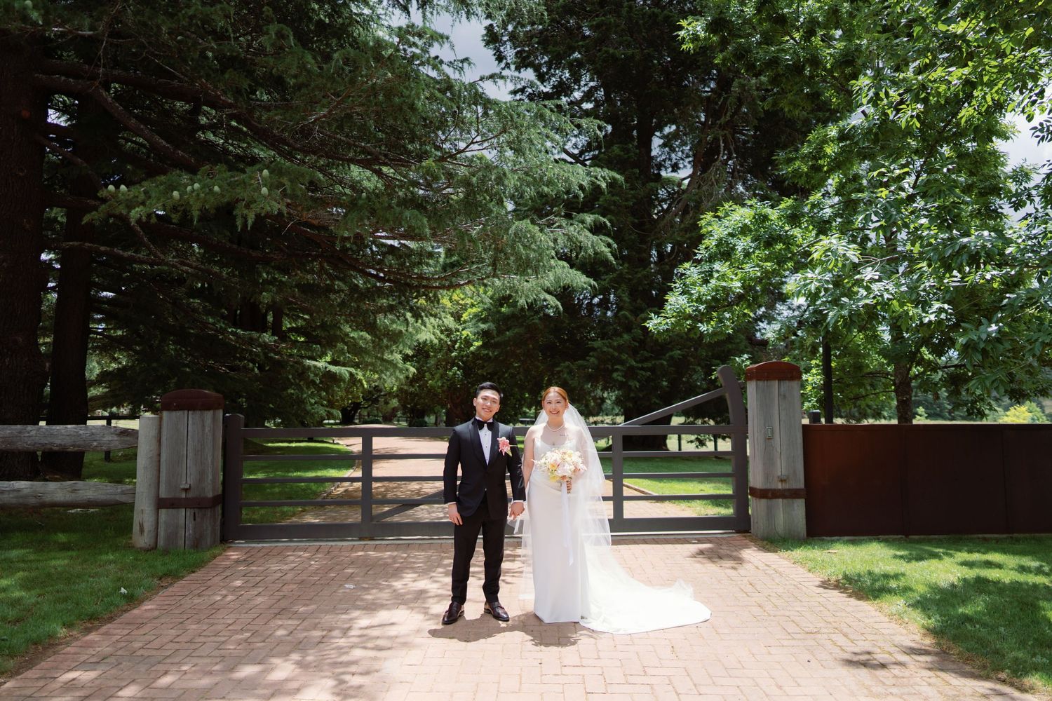 Wedding couple walks along tree-lined path in outdoor garden setting.