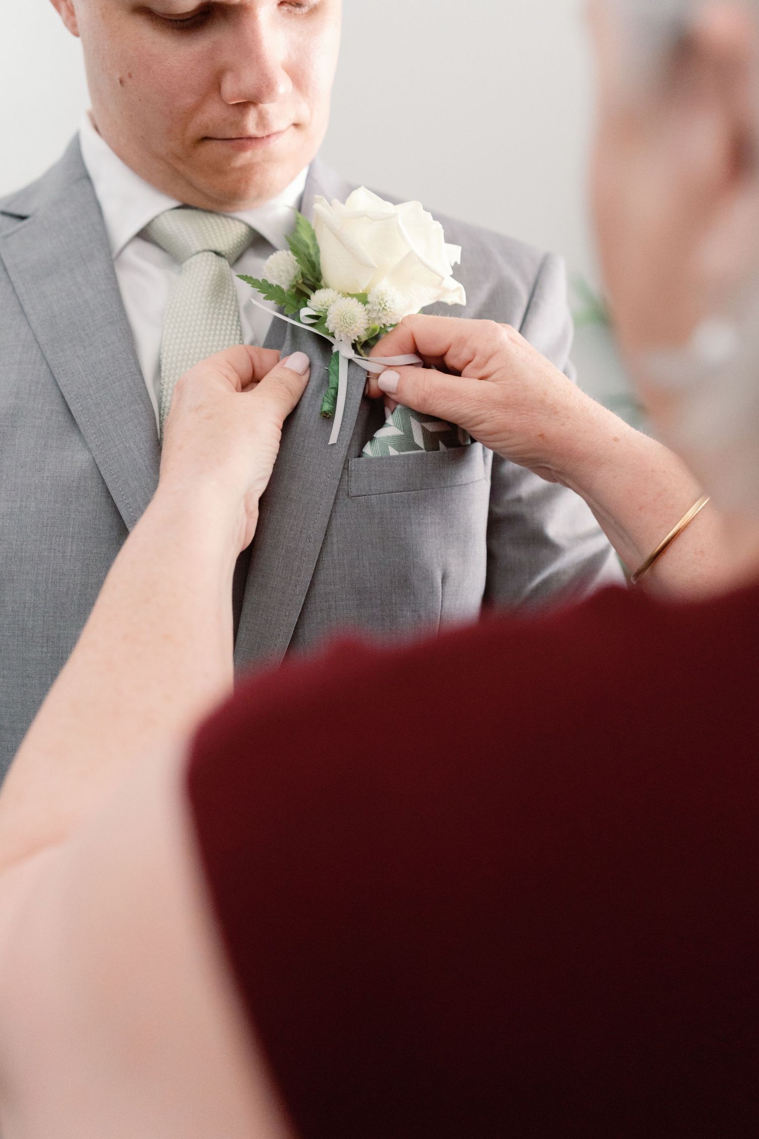 Someone's hands carefully pin a white floral boutonniere onto a gray suit lapel in a wedding preparation sequence.