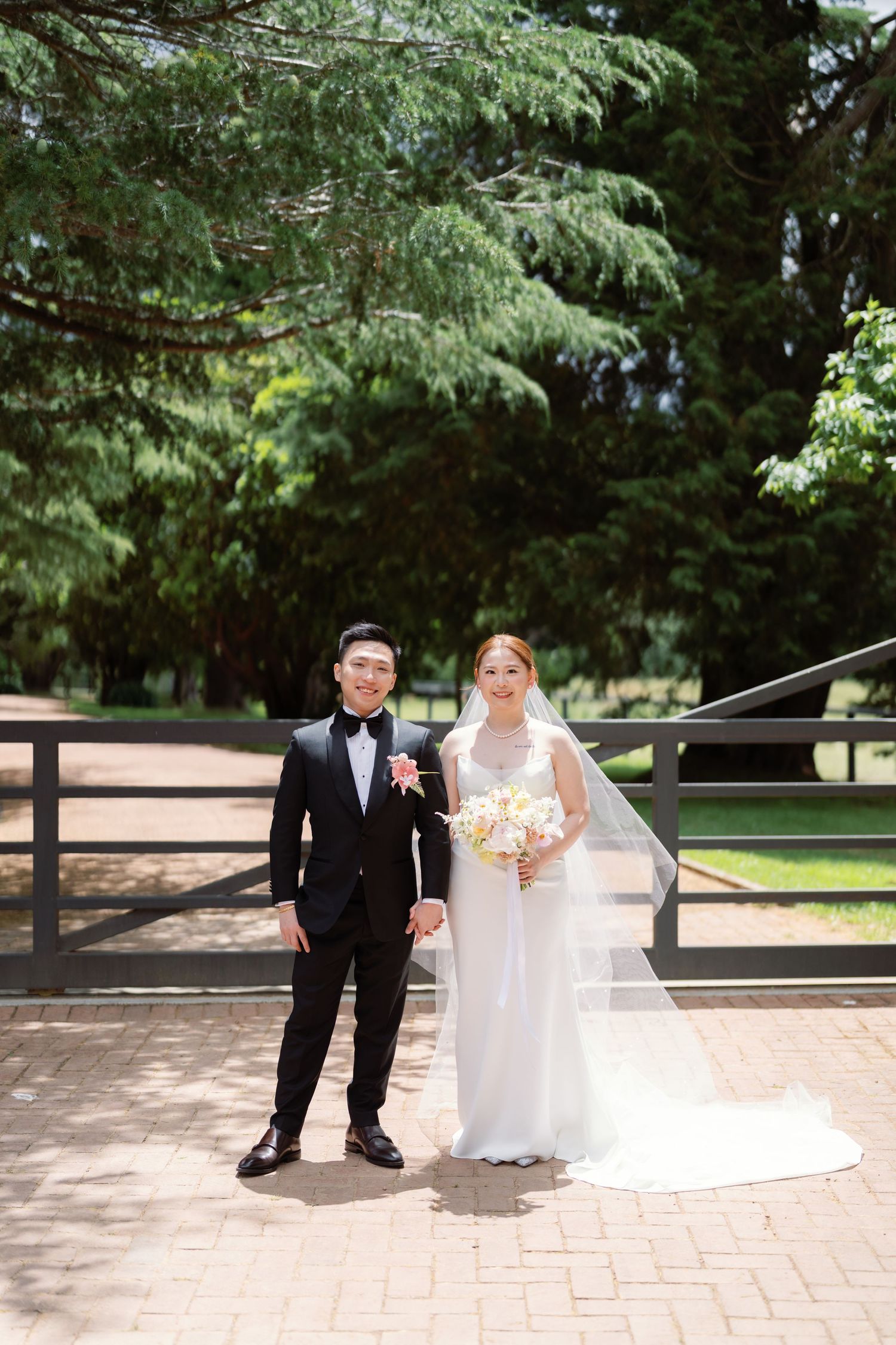 A wedding couple walks together on a brick pathway surrounded by lush green trees in a park setting.