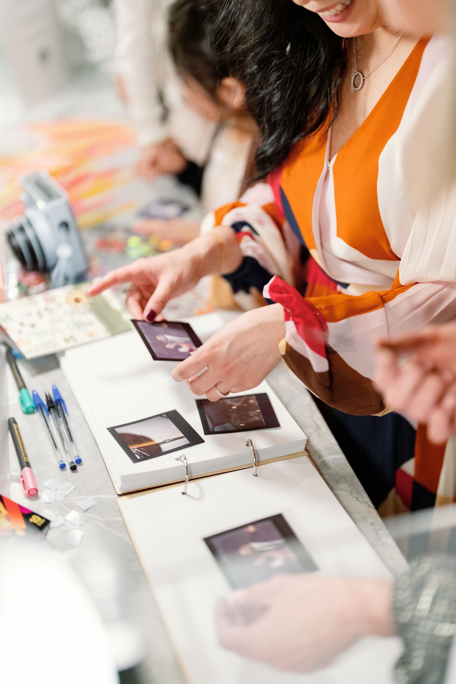 Women reviewing Polaroid photos in a white album on a table with pens and craft supplies nearby.