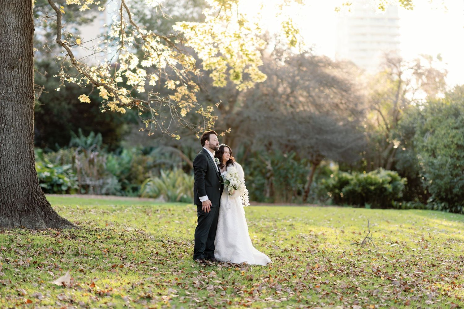 Bride and groom share romantic moment under golden autumn tree in lush garden setting.