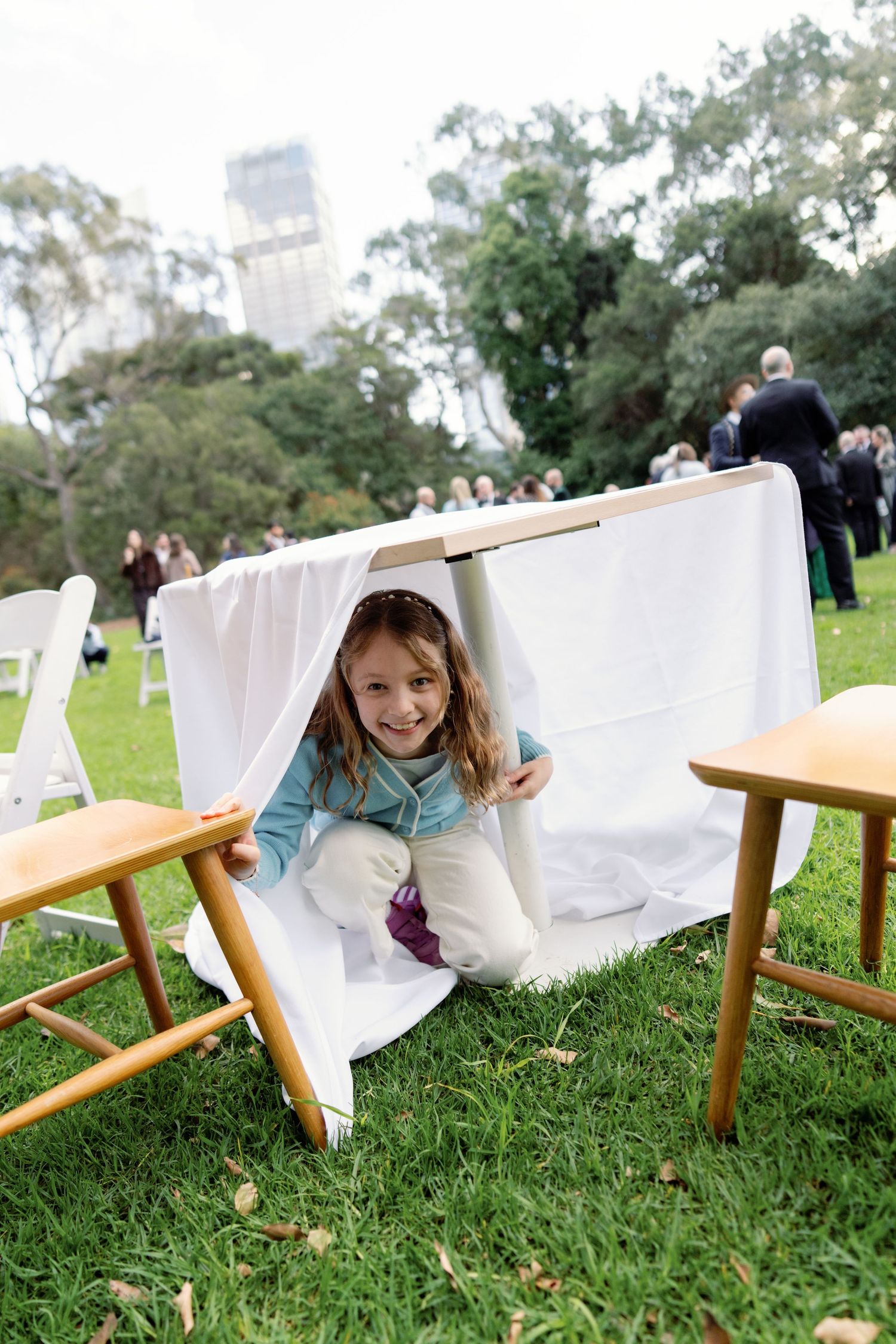 A smiling young girl plays under a white tablecloth at an outdoor wedding reception on green grass.