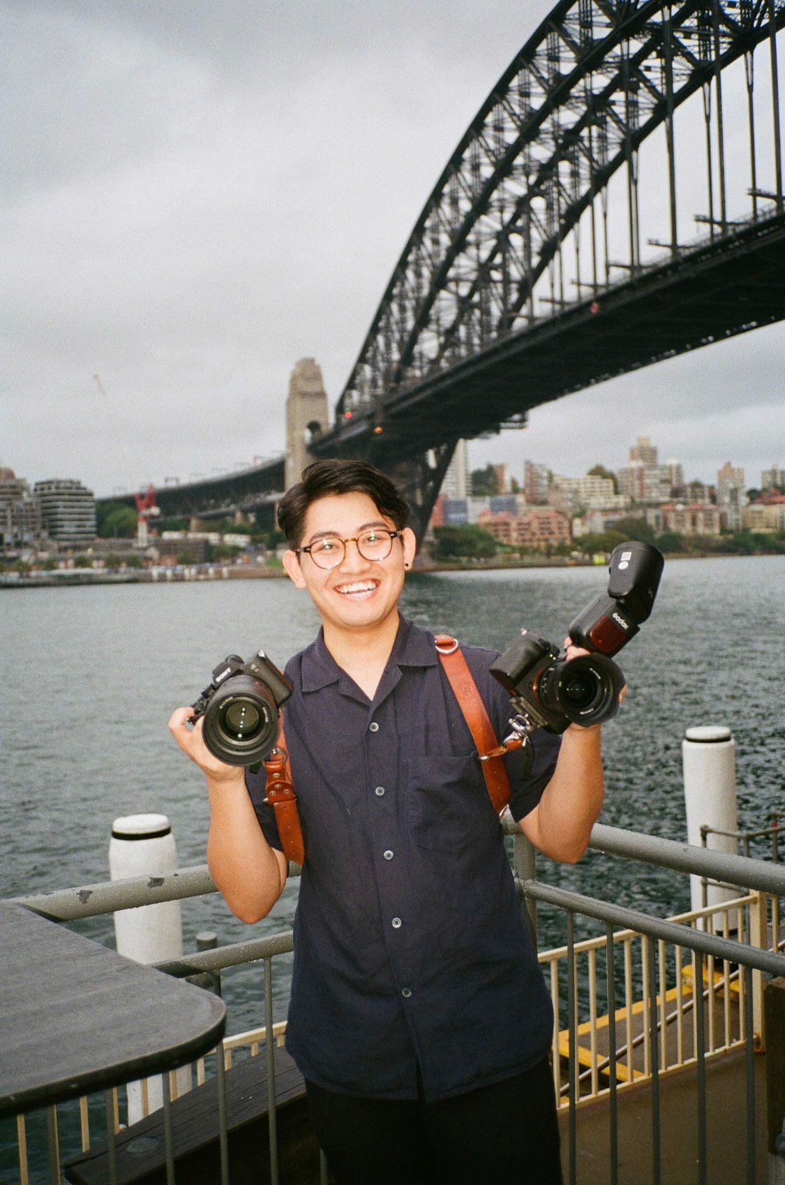 A photographer holding two cameras poses in front of the Sydney Harbour Bridge on an overcast day.