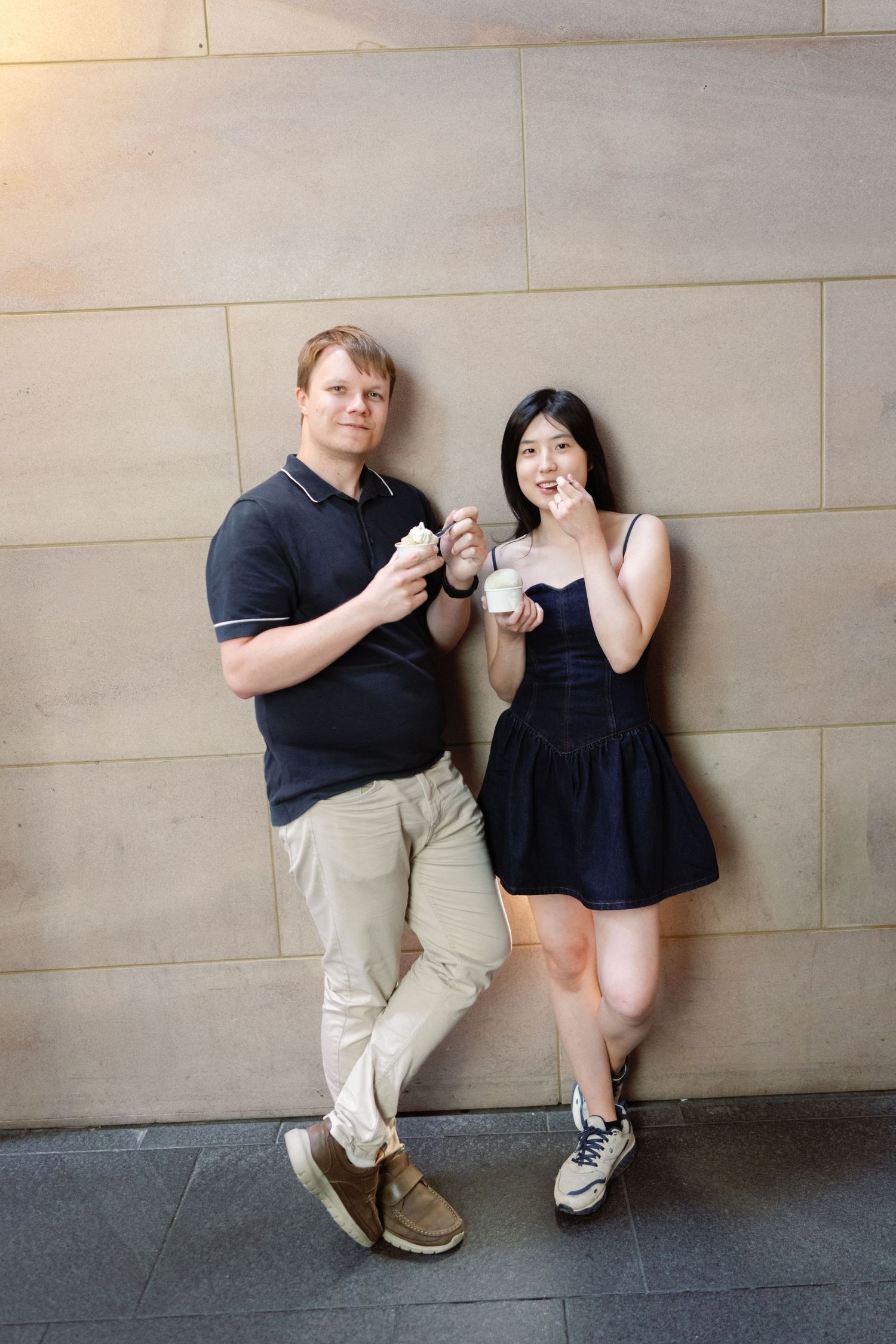 A couple in casual attire stands against a stone wall while enjoying ice cream together.