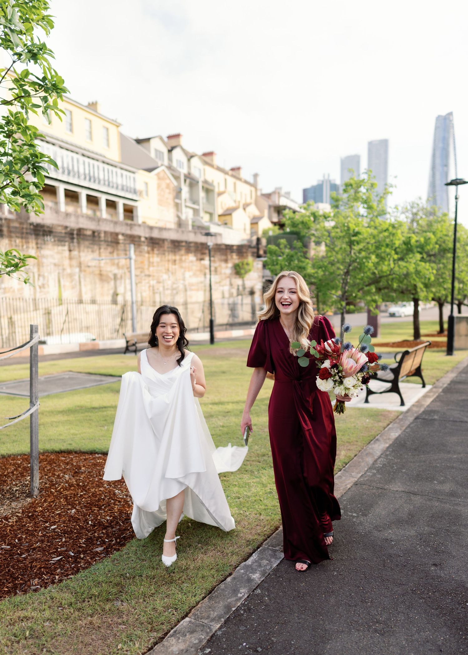 Joyful wedding party poses playfully on path with brick buildings and city skyline in background.