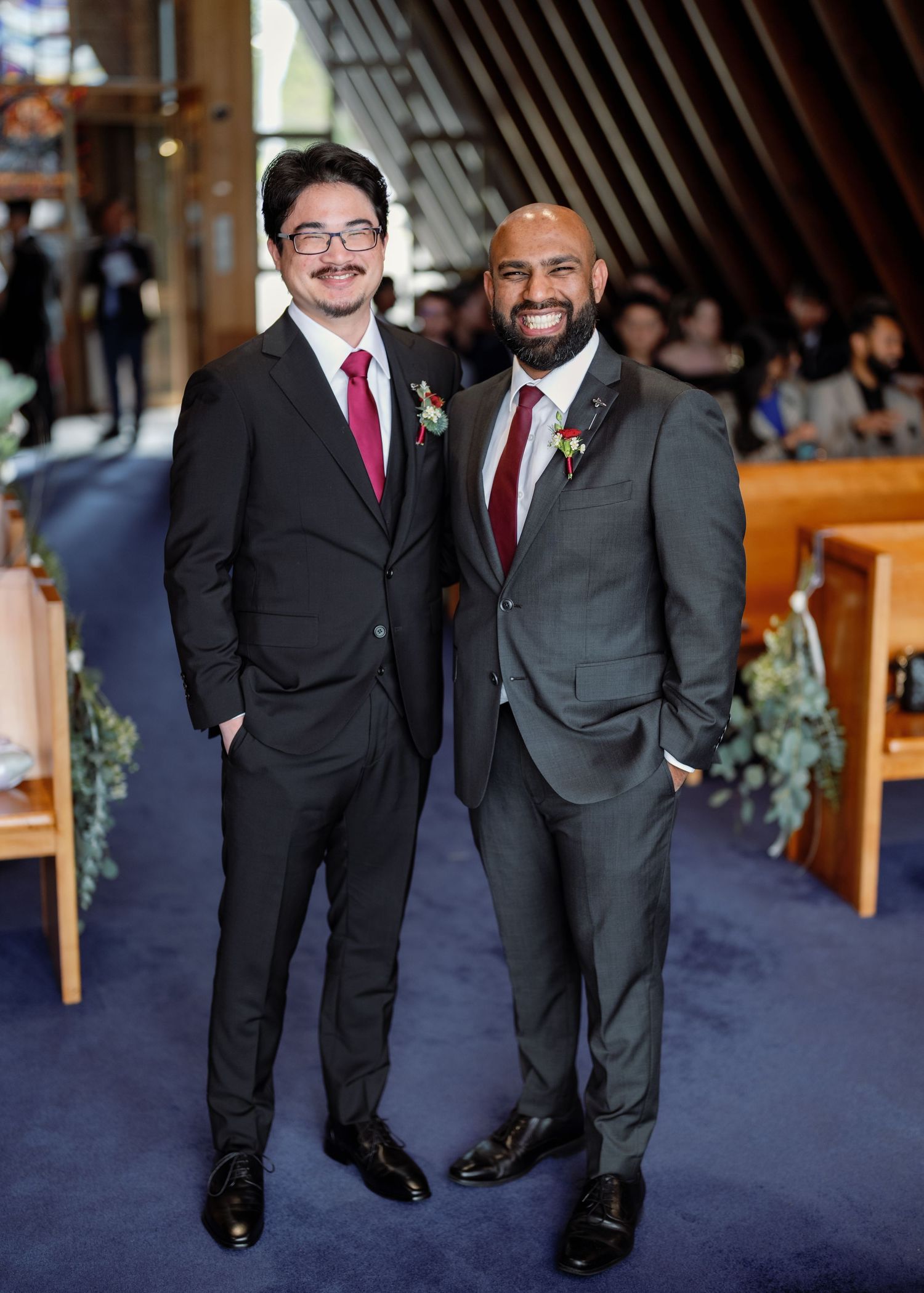 Two people in black suits and burgundy ties stand together on a navy blue carpet at a formal event.