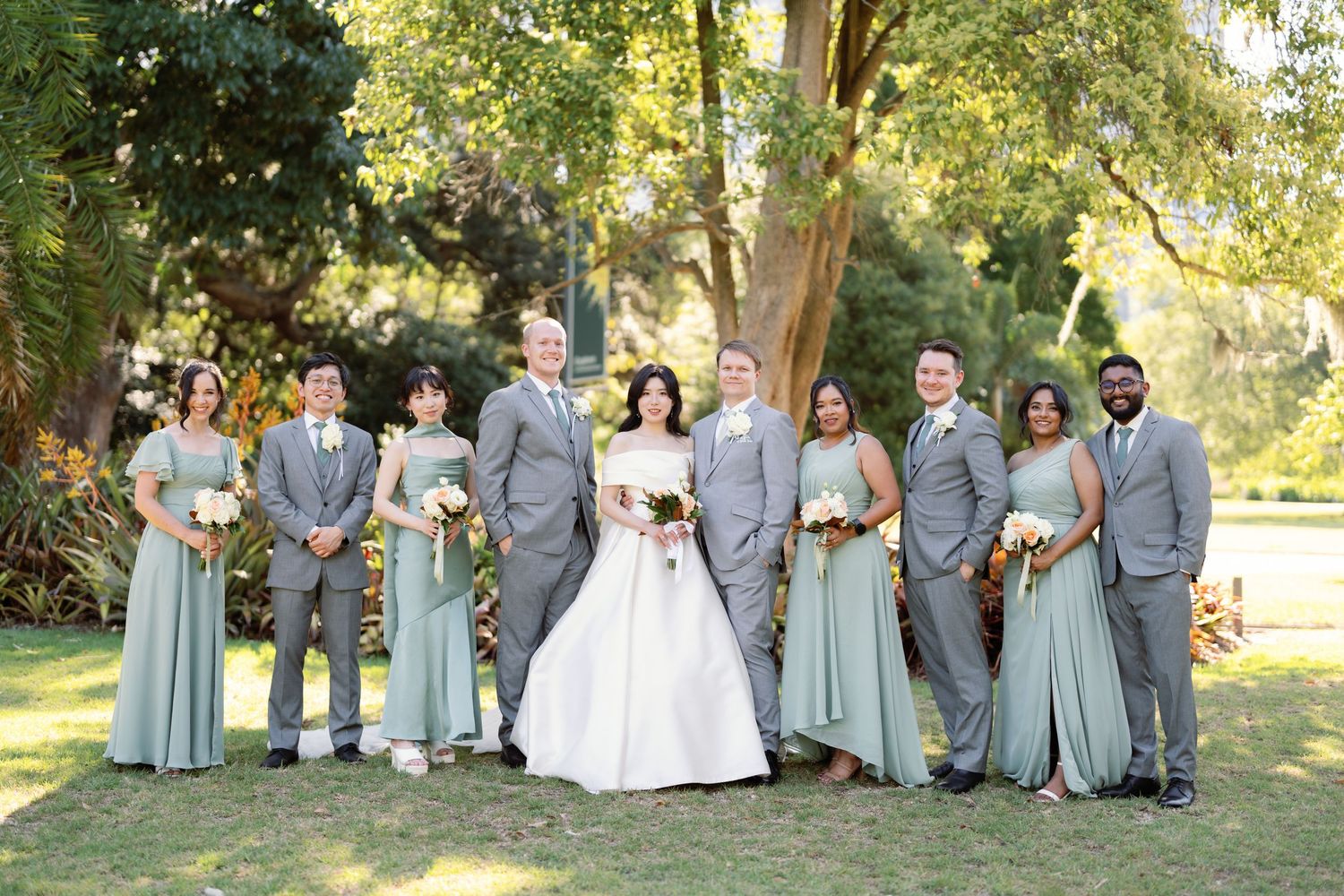 Wedding party in sage green dresses and gray suits poses together outdoors in a natural garden setting.