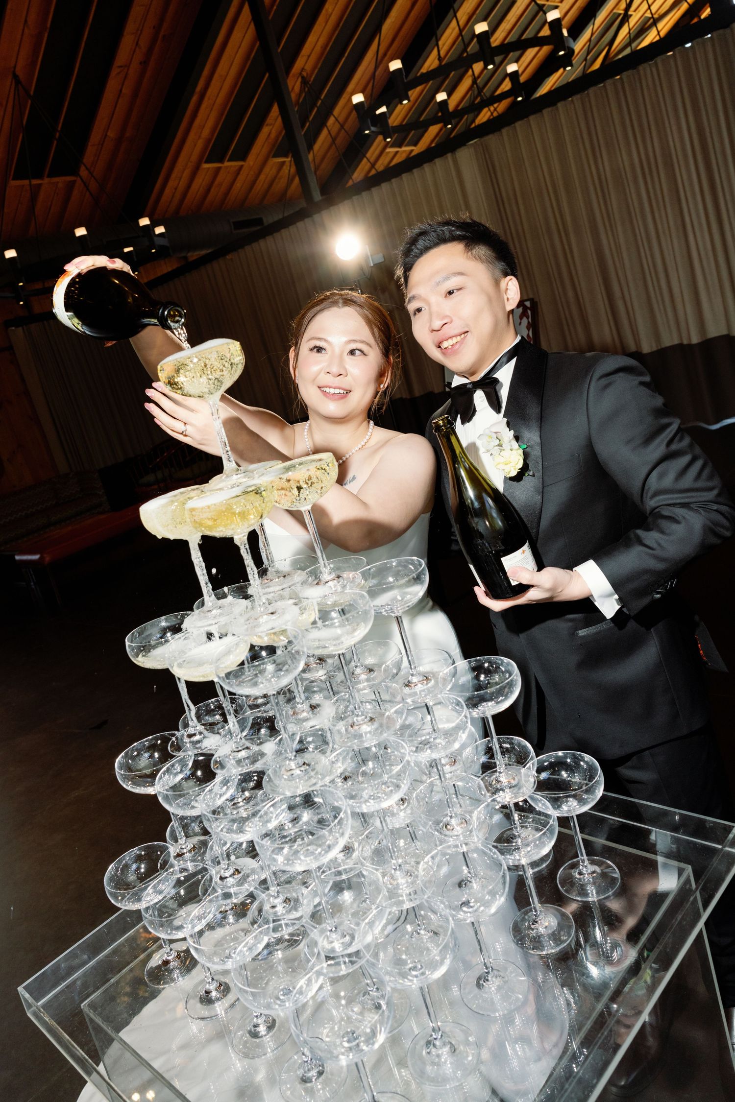 Couple pouring champagne into a tower of crystal glasses at elegant wedding reception.