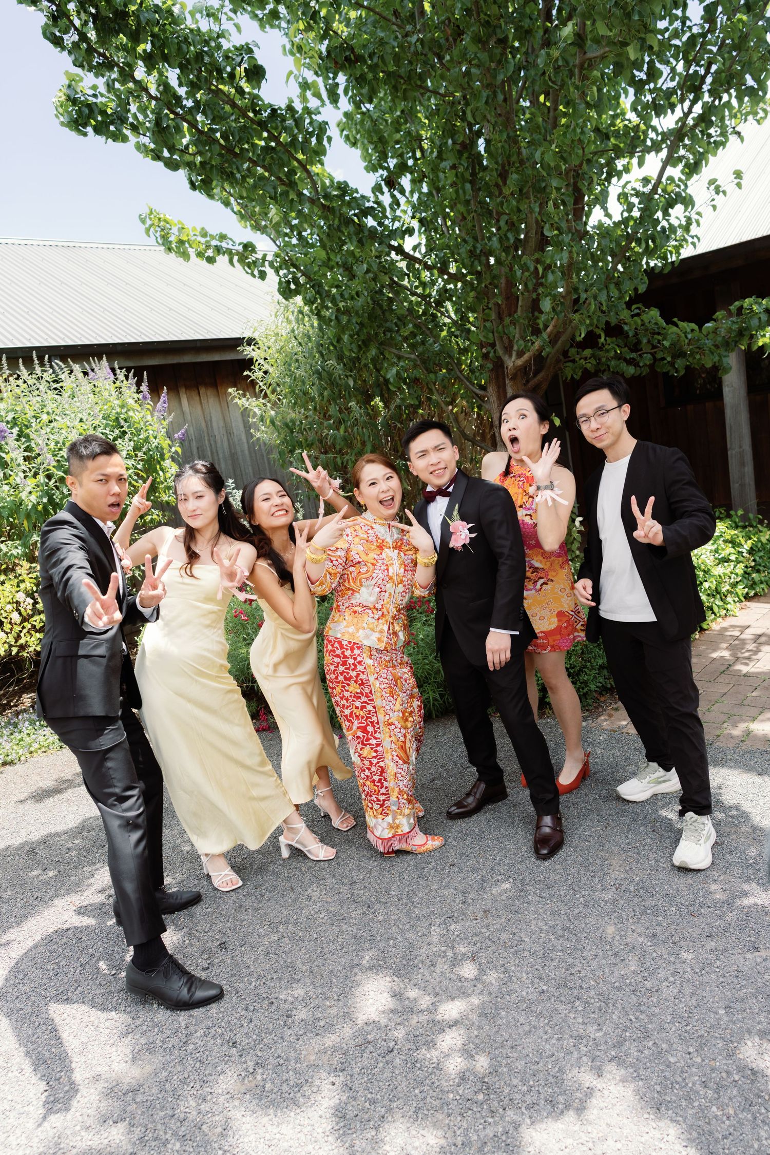 Wedding party poses playfully outdoors under green trees on a sunny day.