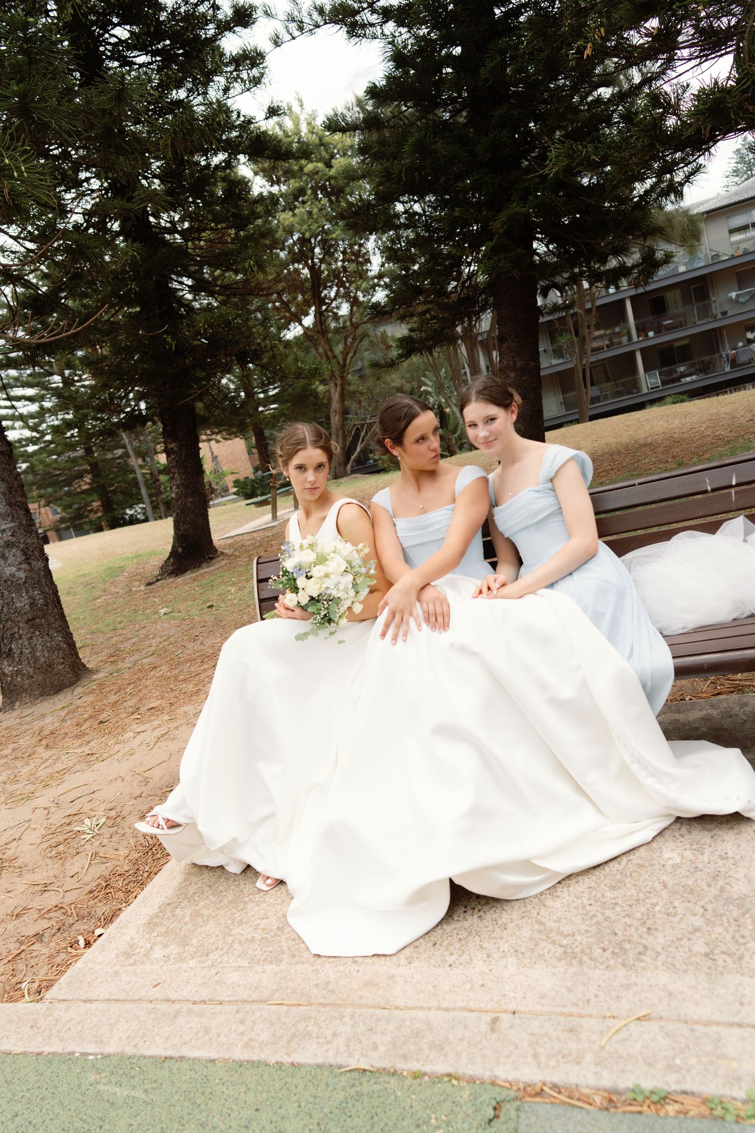 Three women in white and light blue dresses lounging on blanket outdoors.