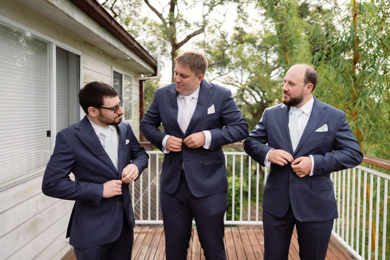 Three groomsmen in matching navy suits share a lighthearted moment on a deck during wedding preparations.