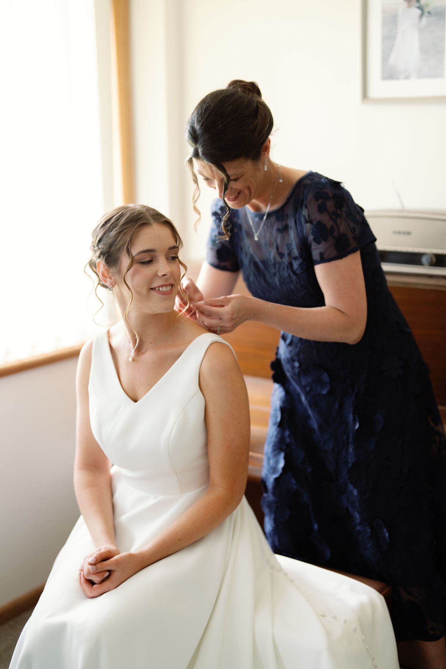 Bride in white wedding dress getting ready with help from attendant in navy blue.
