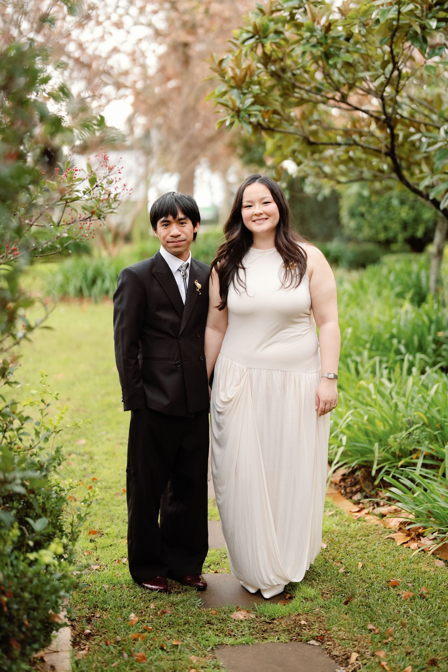 Couple in formal attire posing in a lush garden, man in black suit, woman in elegant white gown.