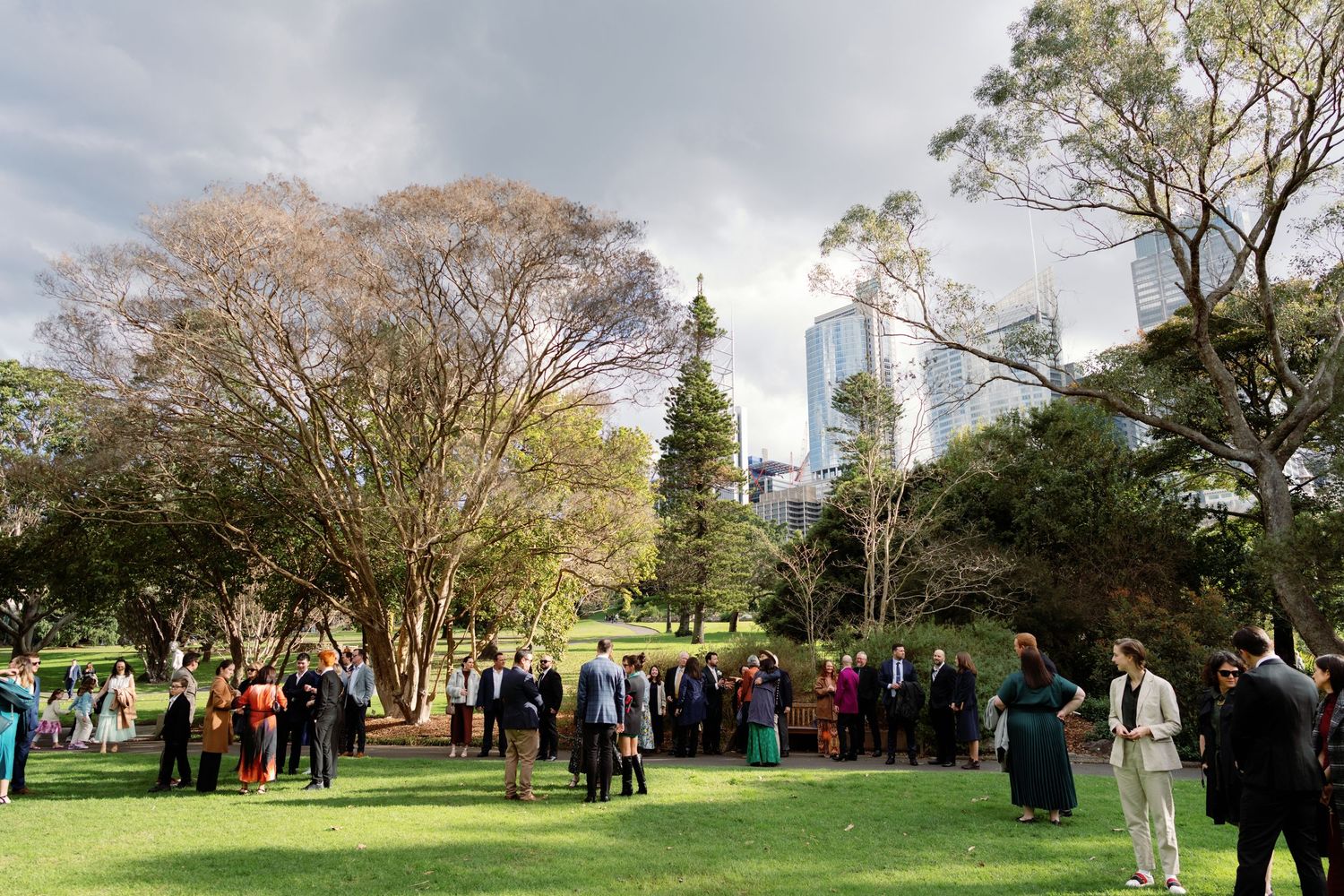 Outdoor wedding reception in a lush park with city skyline, guests mingling on green lawn surrounded by trees.