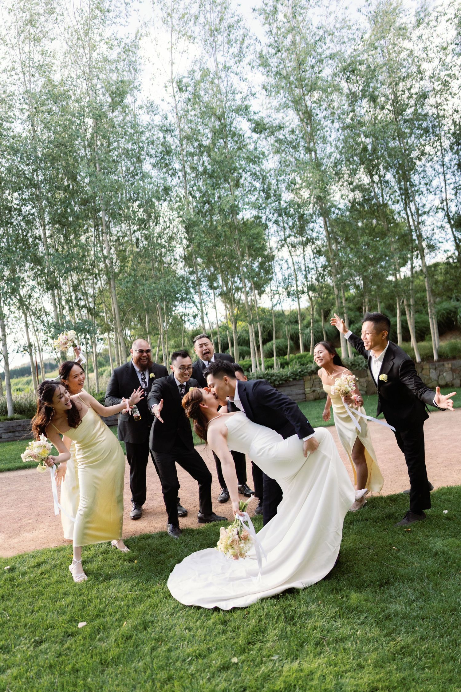 Wedding party in formal attire poses playfully on grassy hill during outdoor celebration.
