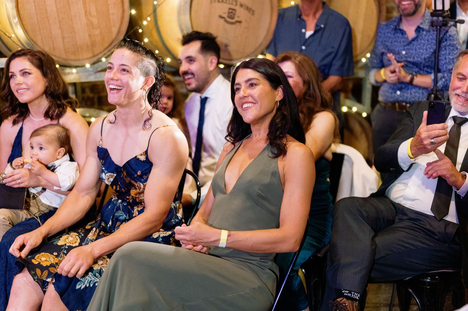 Wedding guests share joyful laughter during a reception celebration with large wine barrels in the background.