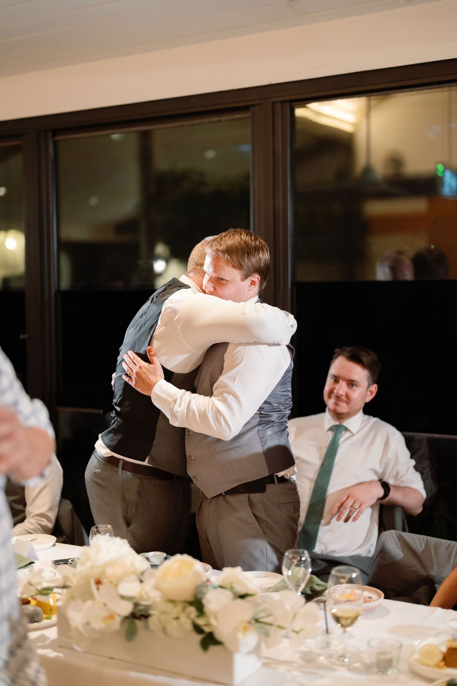 Candid reception moments showing groomsmen hugging and celebrating at a wedding dinner table.